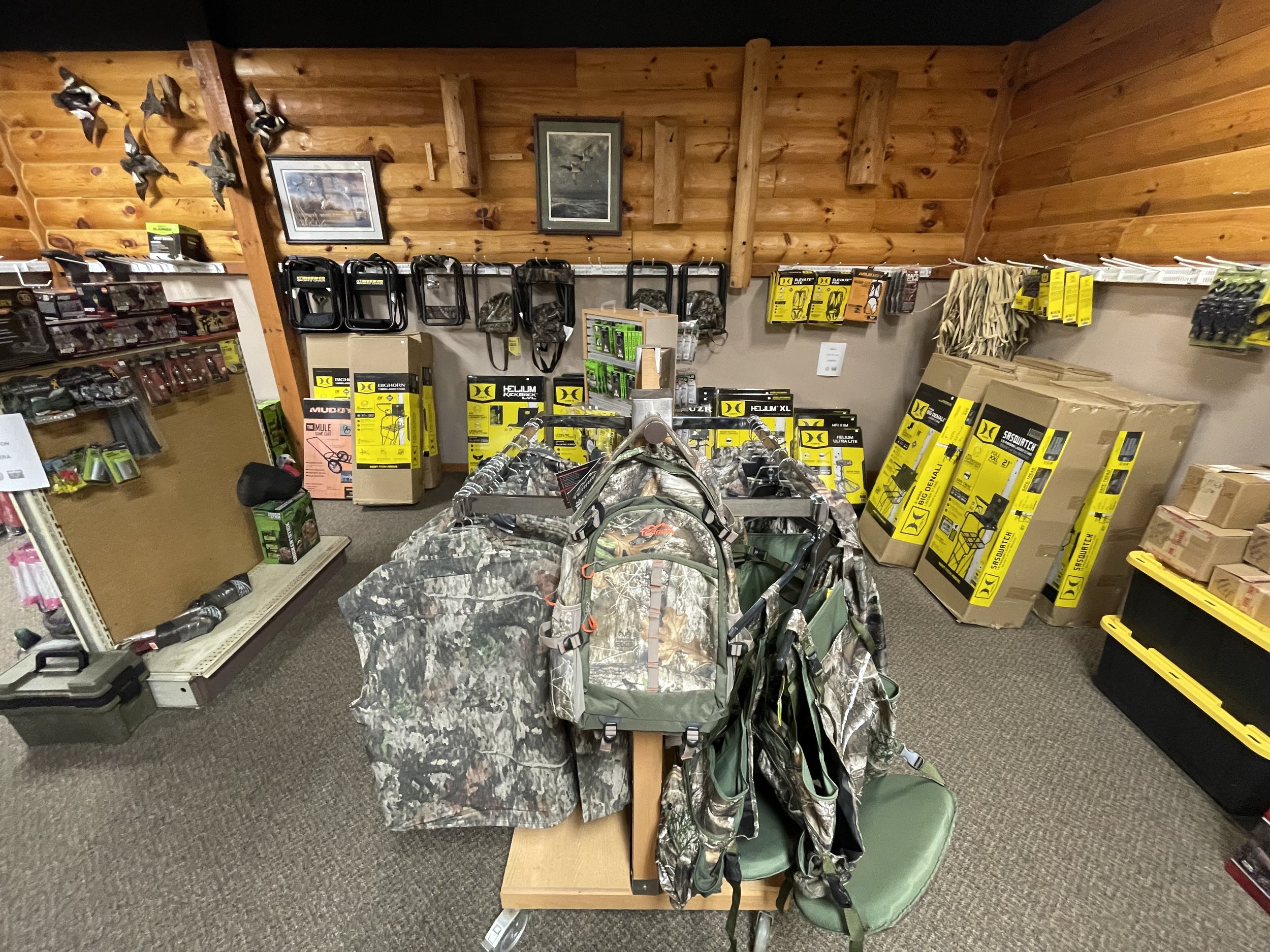 The image shows a store aisle with outdoor gear, including camouflage backpacks, boxes of hunting and outdoor equipment, and black metal chairs hanging on the wooden-paneled wall. There are also framed pictures of birds on the wall.