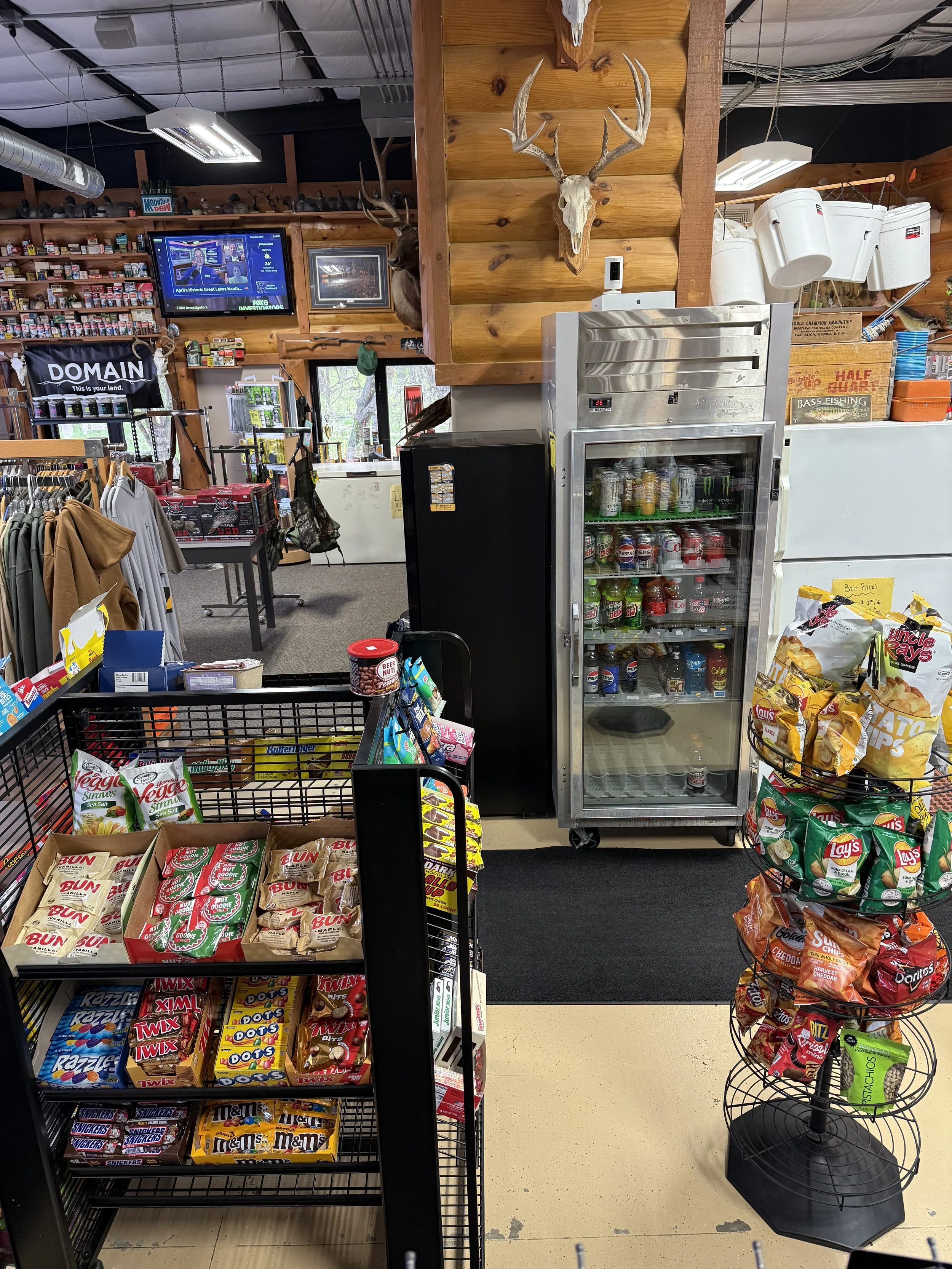 Inside a convenience store with snack shelves, a beverage fridge, a display with snacks, checkout area, and decorative elements like mounted animal antlers and a mounted deer head.