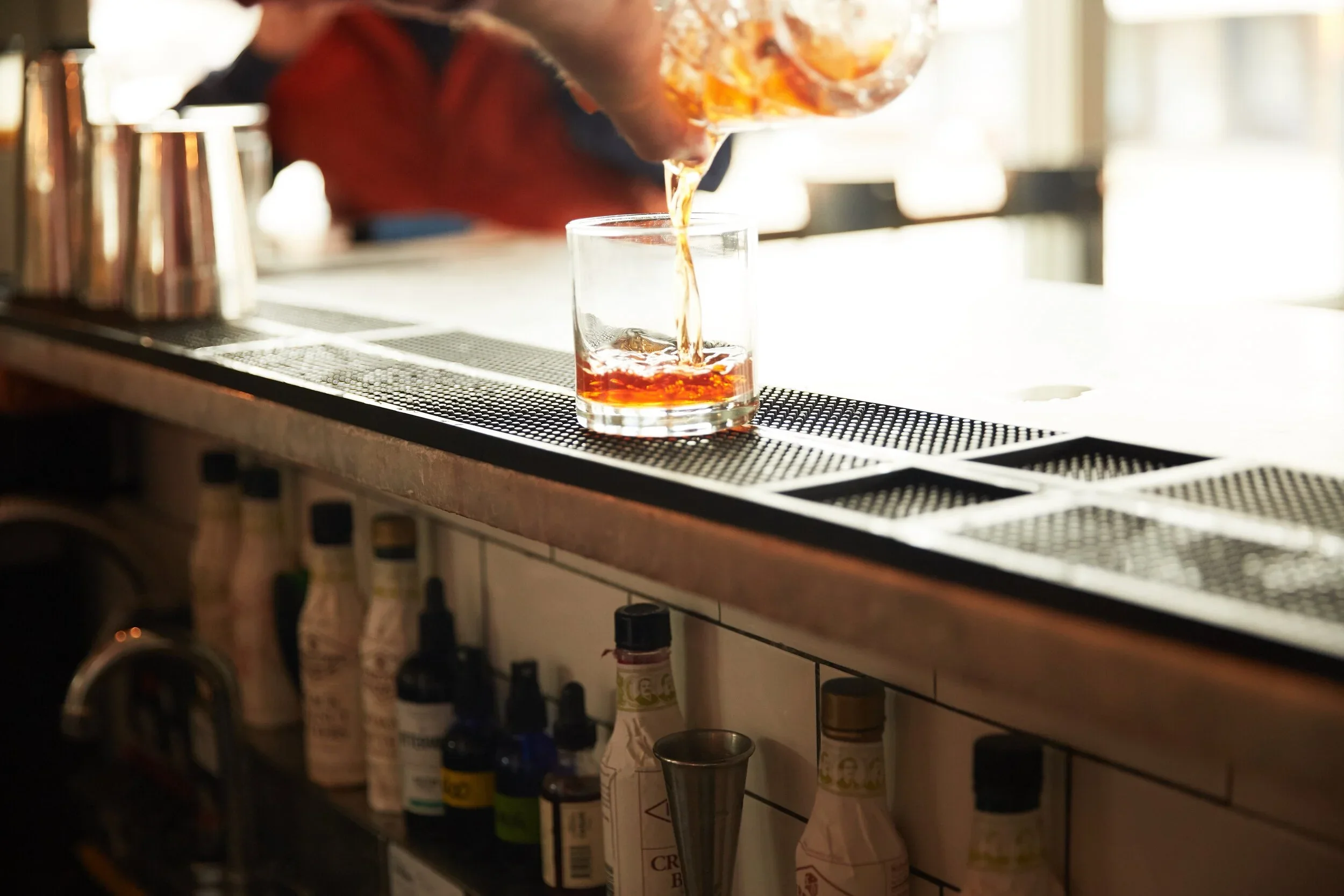 A person pouring amber-colored liquid into a glass at a bar or restaurant setting with various bottles and bar tools in the background.