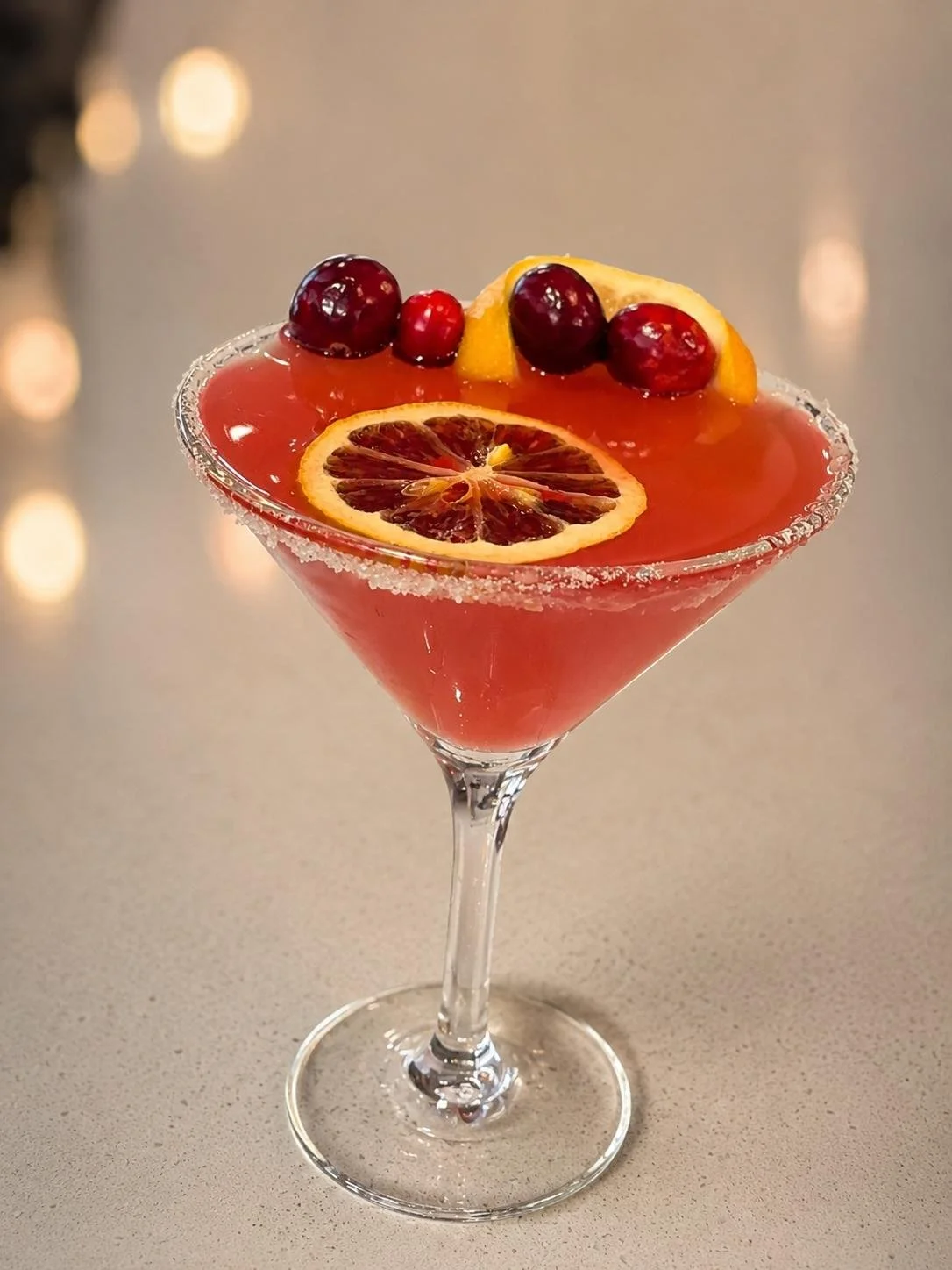 Red cocktail in a martini glass garnished with slices of blood orange and red berries, with a sugared rim, on a gray surface with blurred warm bokeh lights in the background.