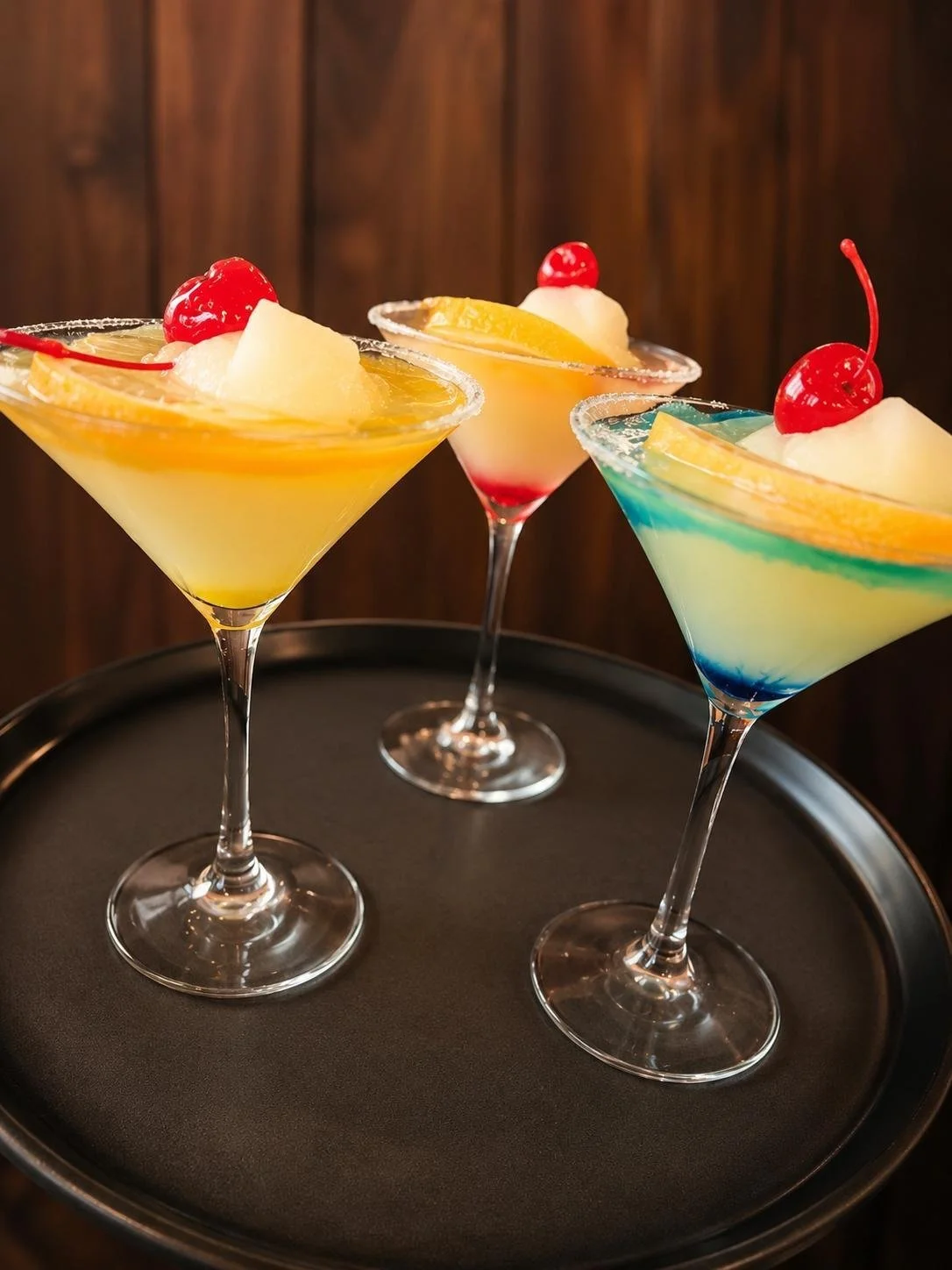 Three colorful cocktails garnished with cherries and fruit slices on a black tray.