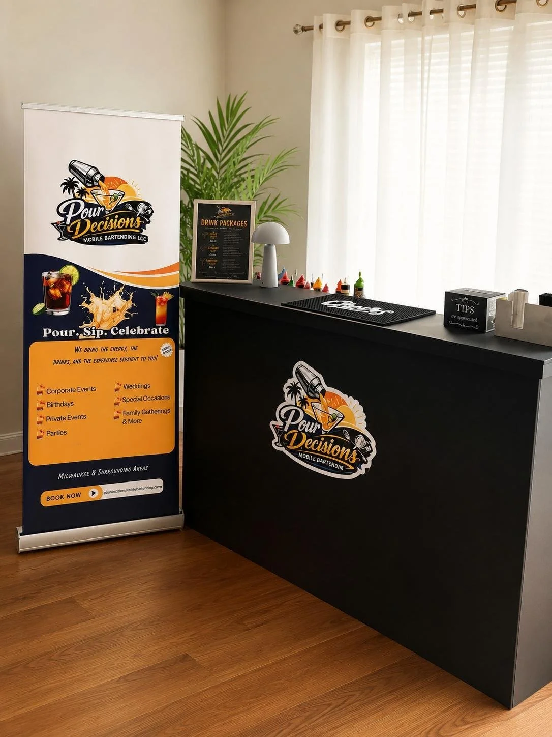 A promotional stand and counter for Pour Decisions Mobile Bartending with drink options, tips box, and colorful liquid coloring bottles, set in a bright room with wood flooring and white curtains.