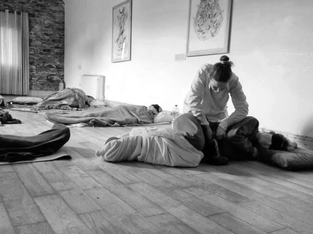 A woman practicing yoga or meditation on a large floor cushion in a room with framed artwork on the wall and sleeping individuals on the floor, some on mats, in the background.