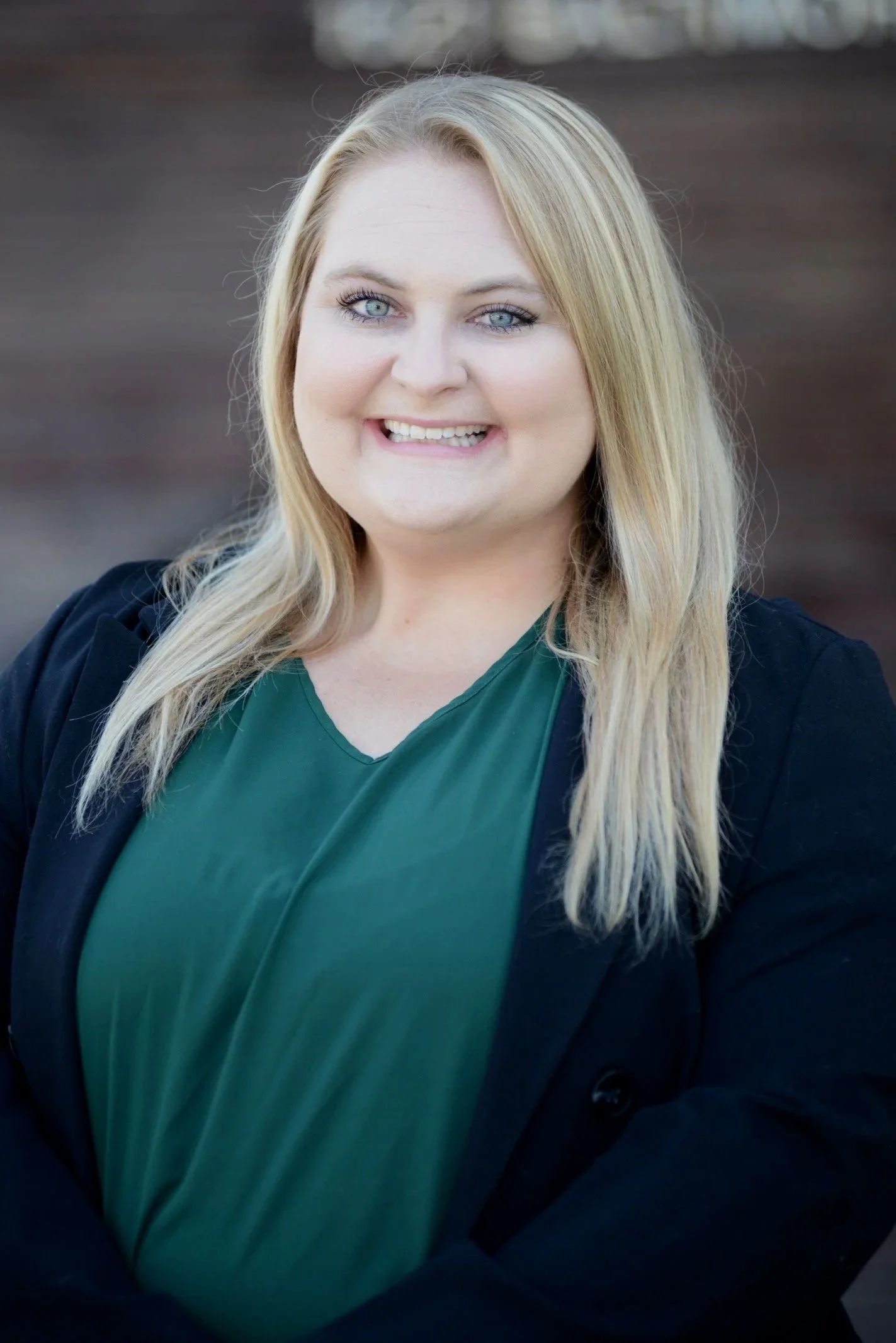 Portrait of a woman with long blonde hair, blue eyes, wearing a dark blazer over a green top, smiling in front of a blurred outdoor background.