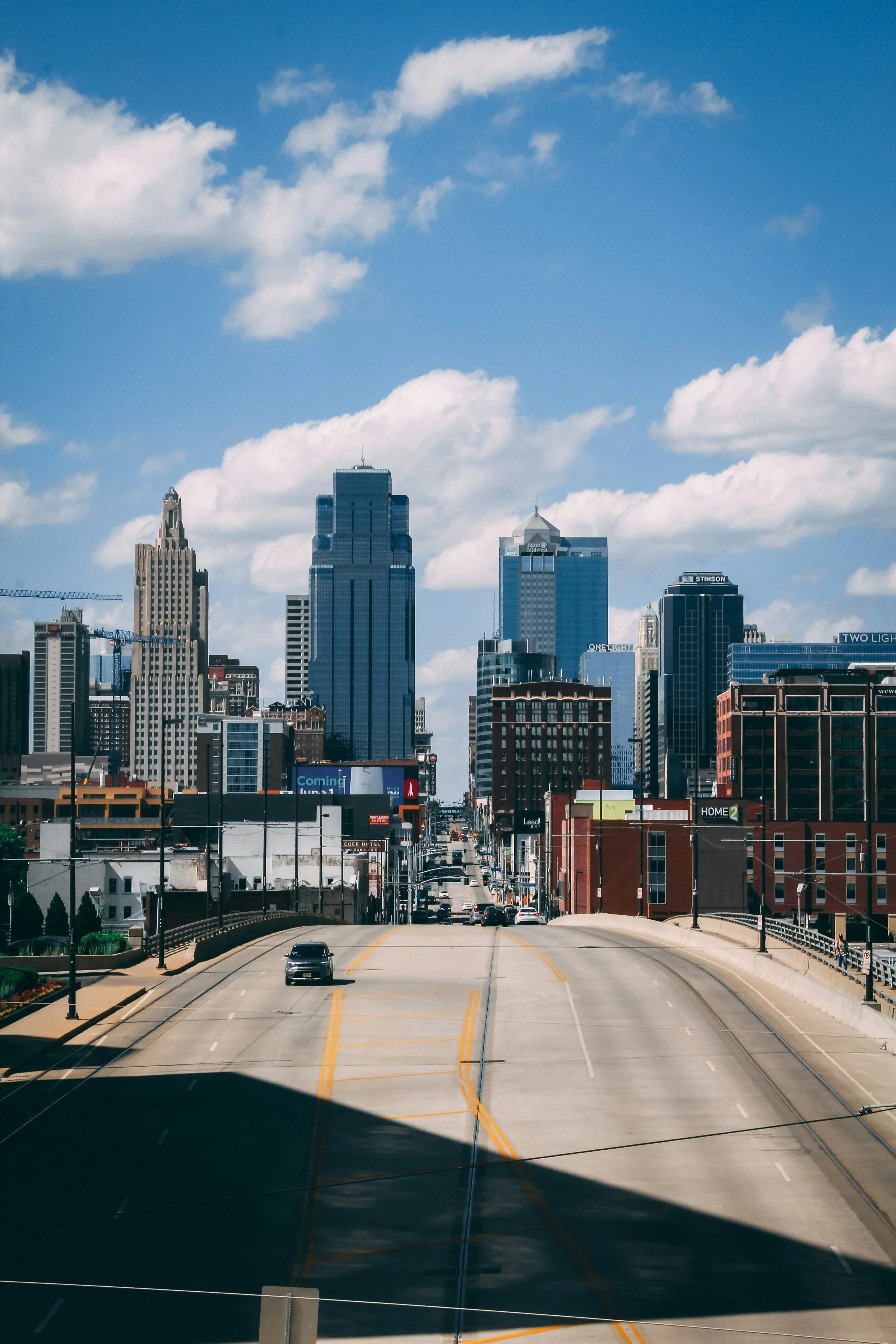 City skyline with tall skyscrapers under a blue sky with scattered white clouds. A bridge or overpass is in the foreground with a few vehicles traveling on the road.