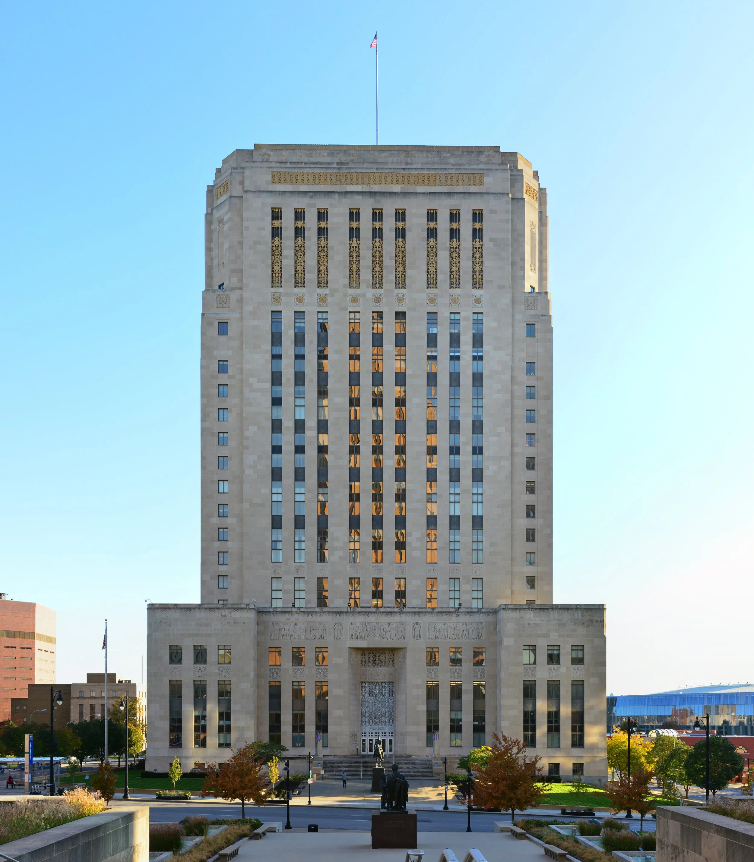 A tall, beige office building with vertical windows and ornate decorative metalwork near the top, with an American flag flying on a flagpole at the roof. In front, there is a small park with trees, sculptures, benches, and walking paths, and blue sky in the background.