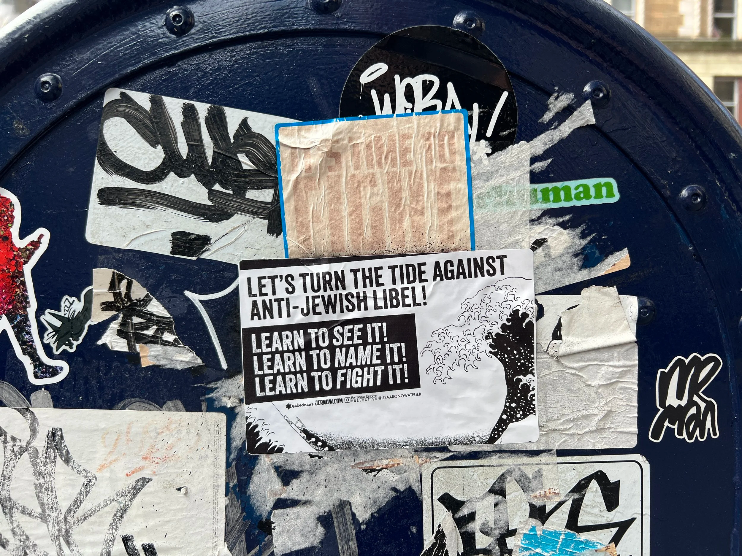 Close-up of a dark blue surface covered with various stickers, including one with bold black text, another with a white background and black text, and a green sticker that says 'man'. The stickers are layered and some are peeling or scratched.