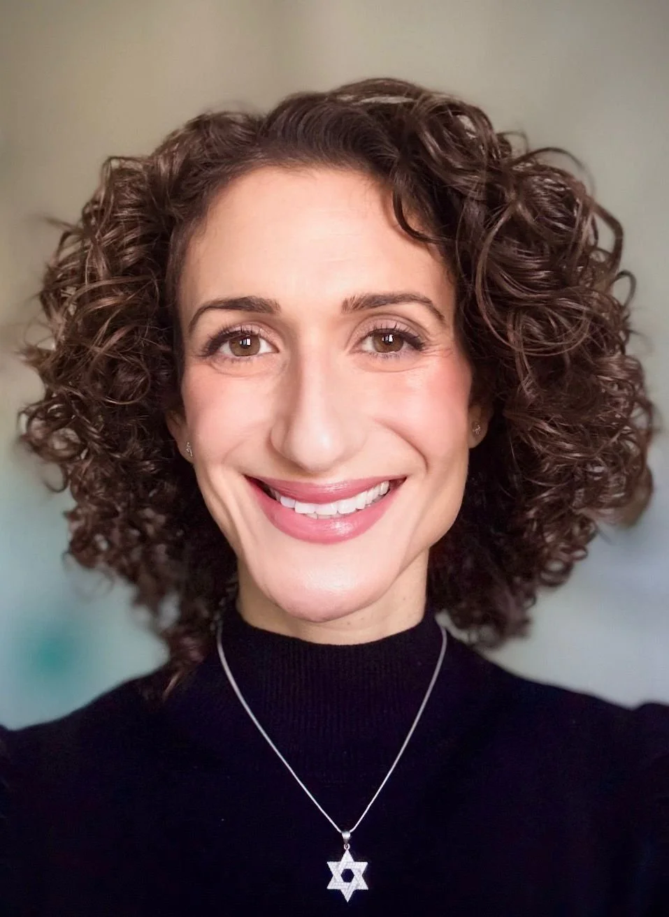 A smiling woman with curly brown hair wearing a black top and a star of David necklace.