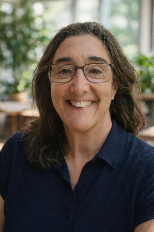 Portrait of a woman with glasses smiling in a bright, indoor setting with green plants in the background.