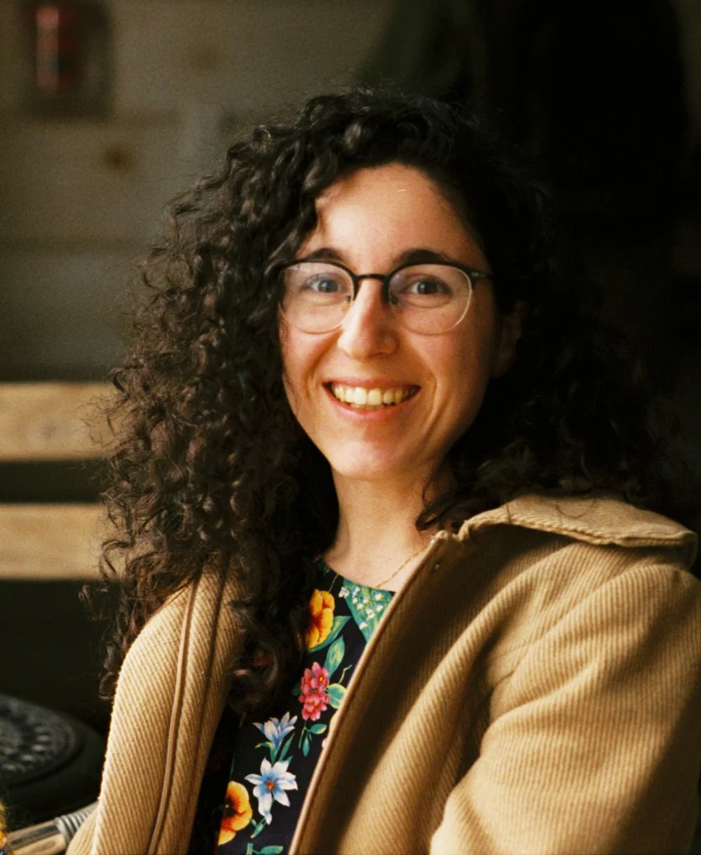 A smiling woman with curly dark hair wearing glasses, a floral blouse, and a tan jacket