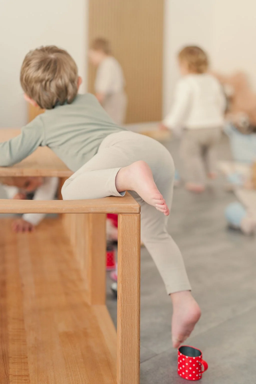 Ein Kind sitzt auf einem Pikler Hozgerüst, mit ausgestrecktem Bein, blickt in die Ferne, während andere Kinder im Hintergrund auf dem Boden spielen.