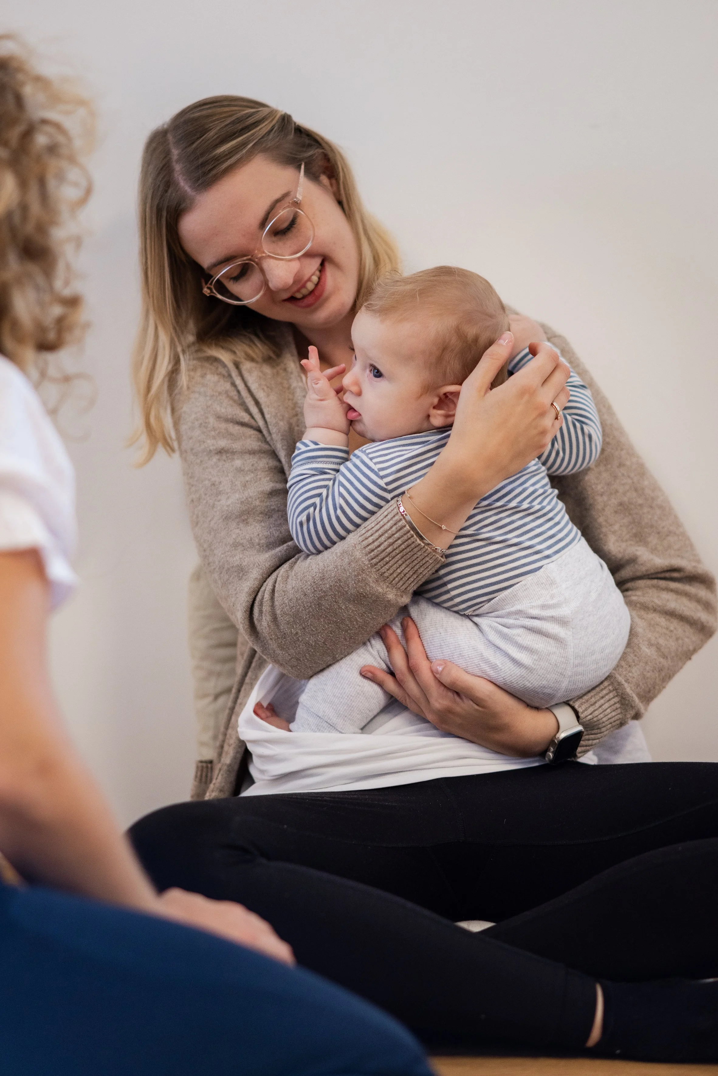 Eine Frau mit Brille hält ein weinendes Baby, das eine Hand auf ihr Gesicht legt, liebevoll im Arm. Weitere Personen sind im Vordergrund und im Hintergrund zu sehen.