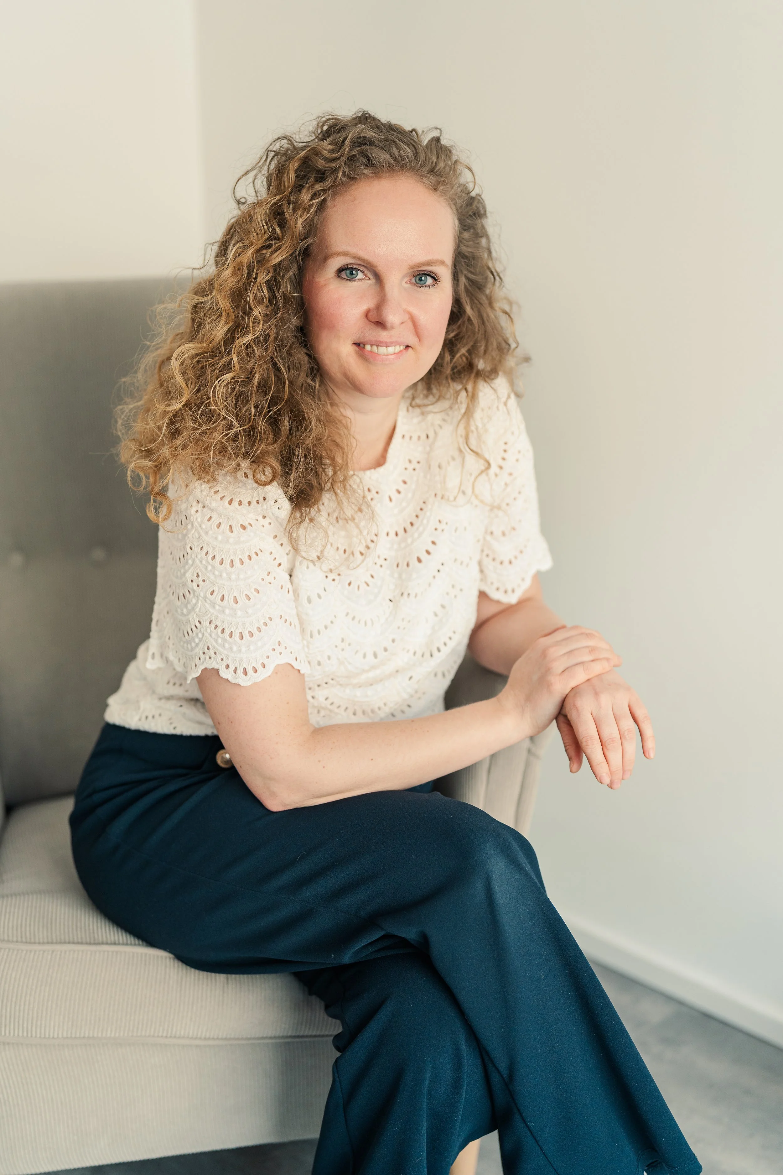 Eine Frau mit lockigem, braunem Haar sitzt auf einem grauen Sofa, trägt eine weiße, lockere Bluse und dunkle Hosen, lächelt und schaut in die Kamera.