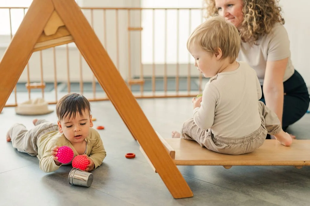 Zwei kleine Kinder spielen auf dem Boden eines Spielzimmers, ein Mädchen liegt auf dem Bauch und hält zwei pinke Spielsachen, während ein Junge auf einer Bank sitzt und von einer Erwachsenenperson, vermutlich einer Erzieherin, beobachtet wird.