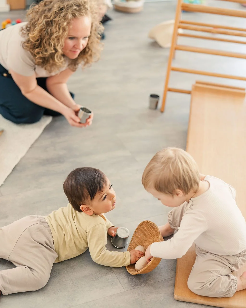 Zwei Kinder auf dem Boden, eines reckt die Hand nach einem Korb aus. Eine erwachsene Frau beobachtet das Geschehen.