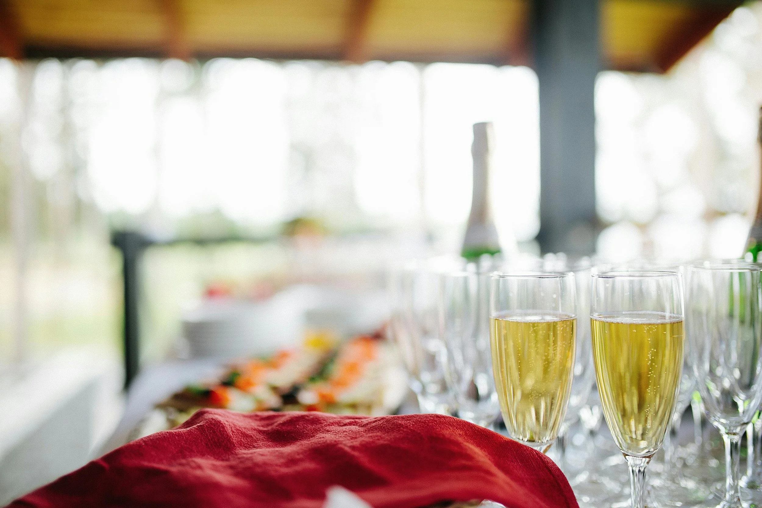 Several glasses filled with a yellow beverage on a table with a red cloth, blurred background with bright light and out-of-focus objects.