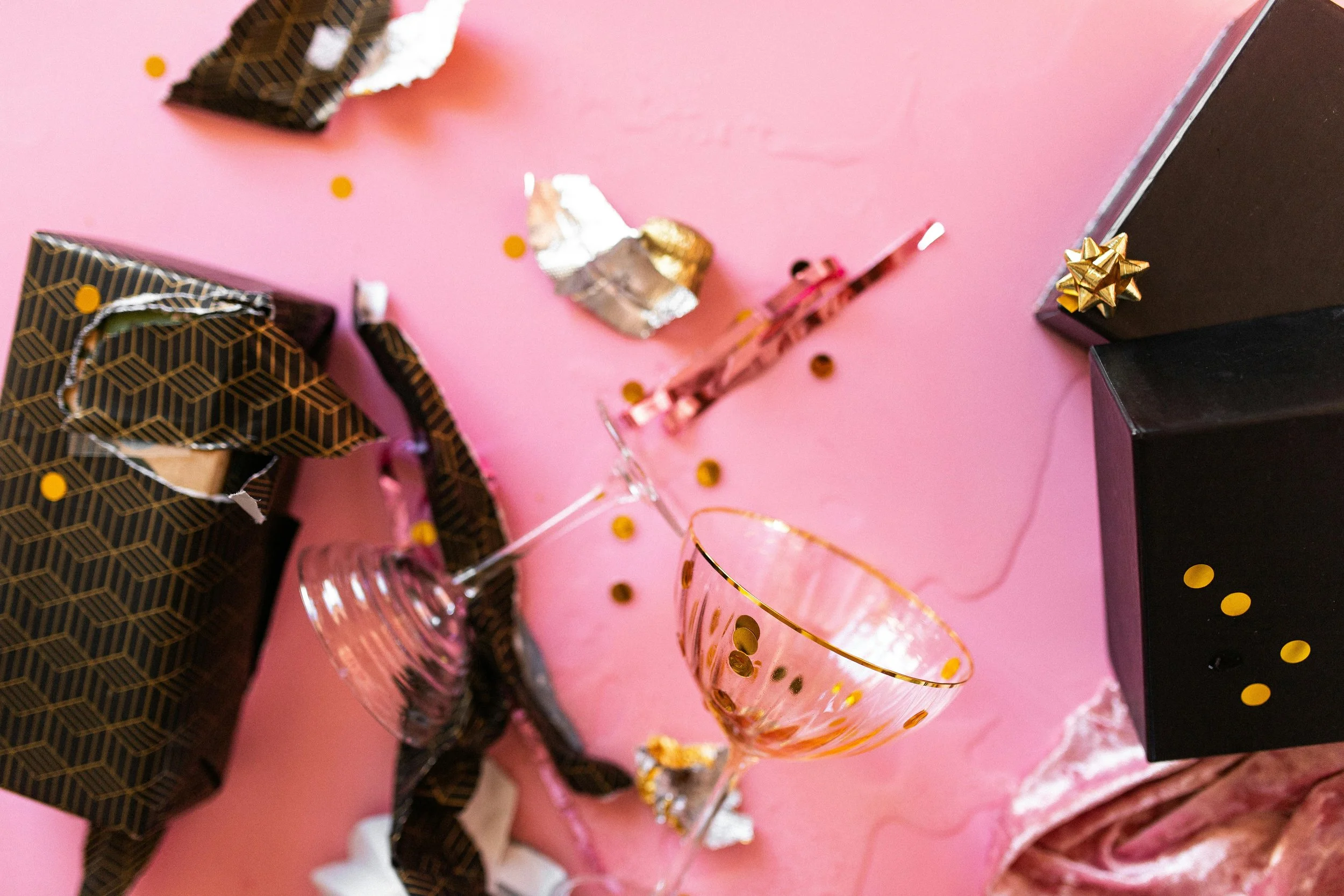 Pink table with torn wrapping paper, gold confetti, and a champagne flute.