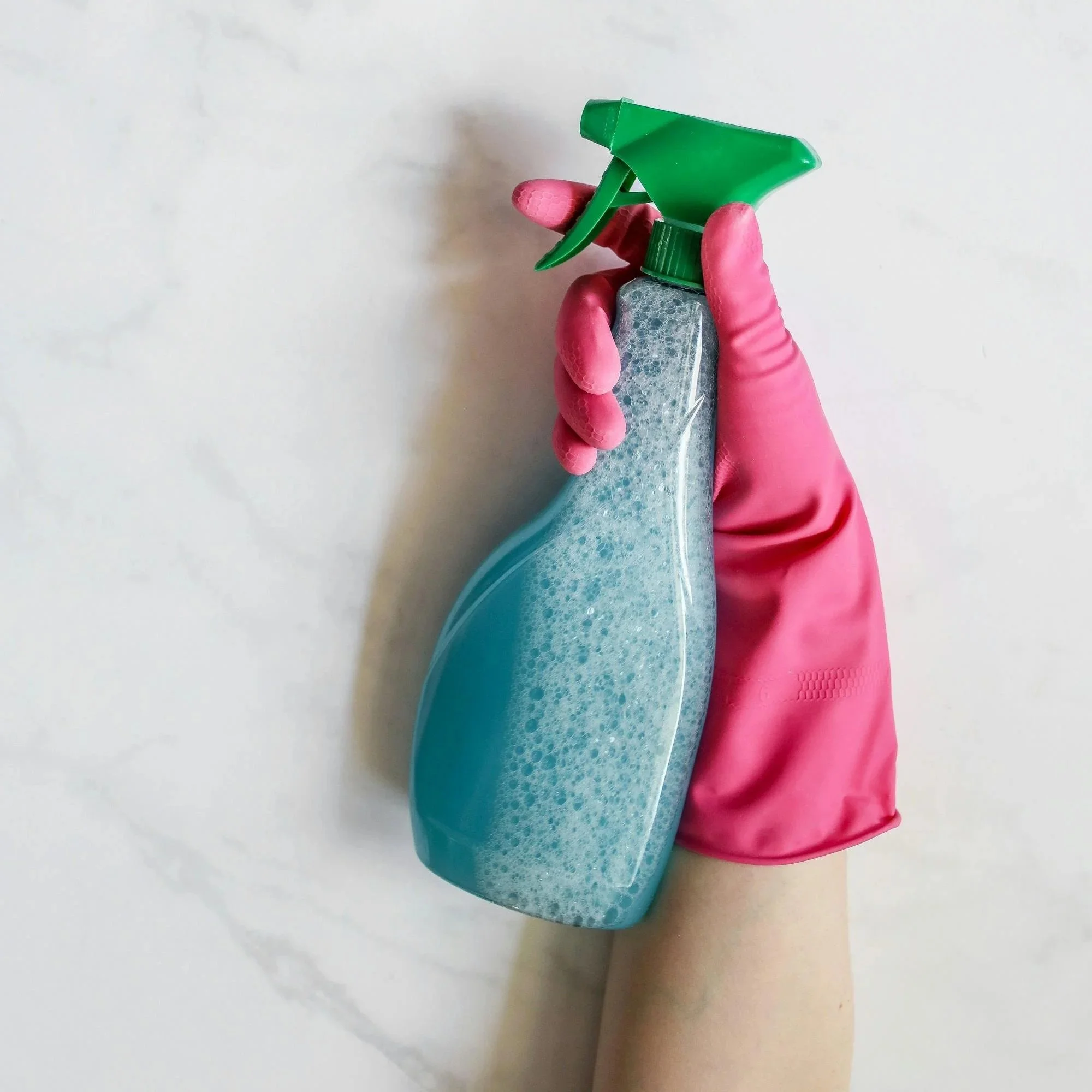 Person holding a spray bottle filled with cleaning liquid, wearing pink rubber gloves, against a white background.