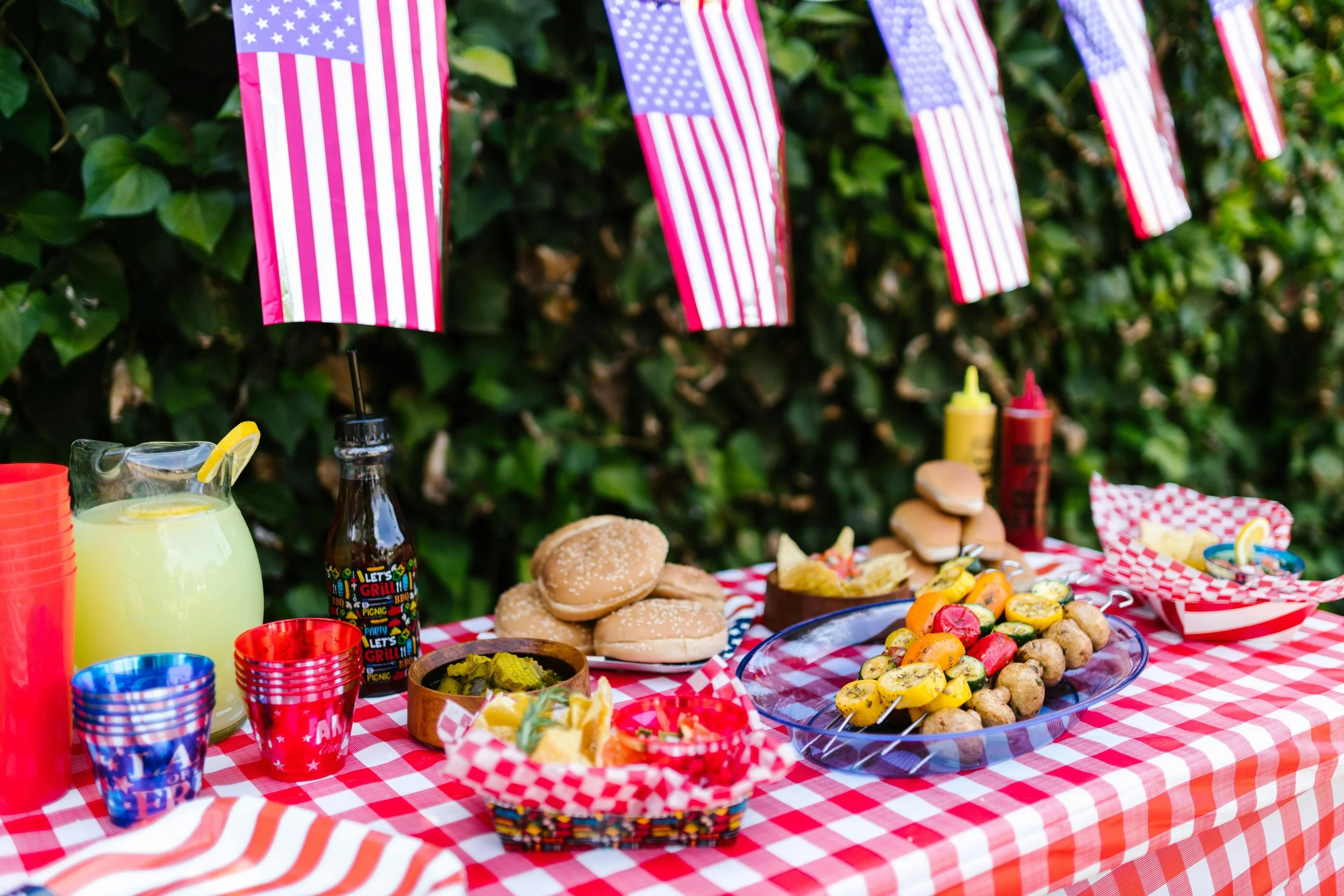 Decorative outdoor table set for a celebration with American flags, colorful cups, lemonade, and a variety of foods including sliders, potato salad, grilled vegetables, and sliced bread, with a green leafy background.