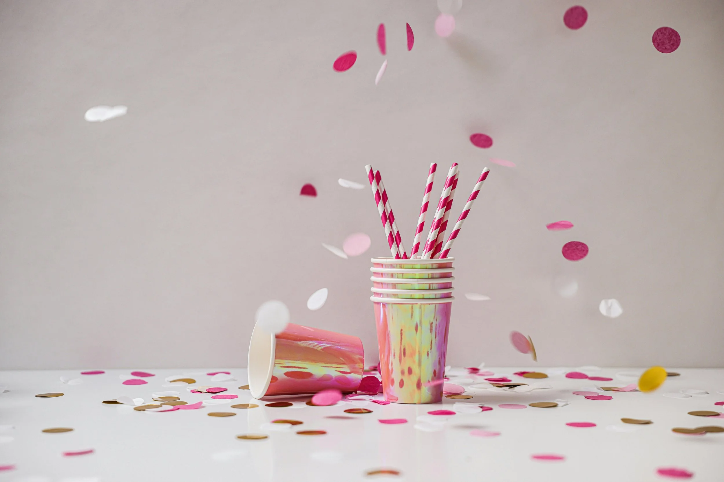 Pink and white striped paper straws in a pastel-colored disposable cup with splashy paint design, surrounded by scattered pink, white, and gold confetti, with some confetti falling from above against a plain gray background.