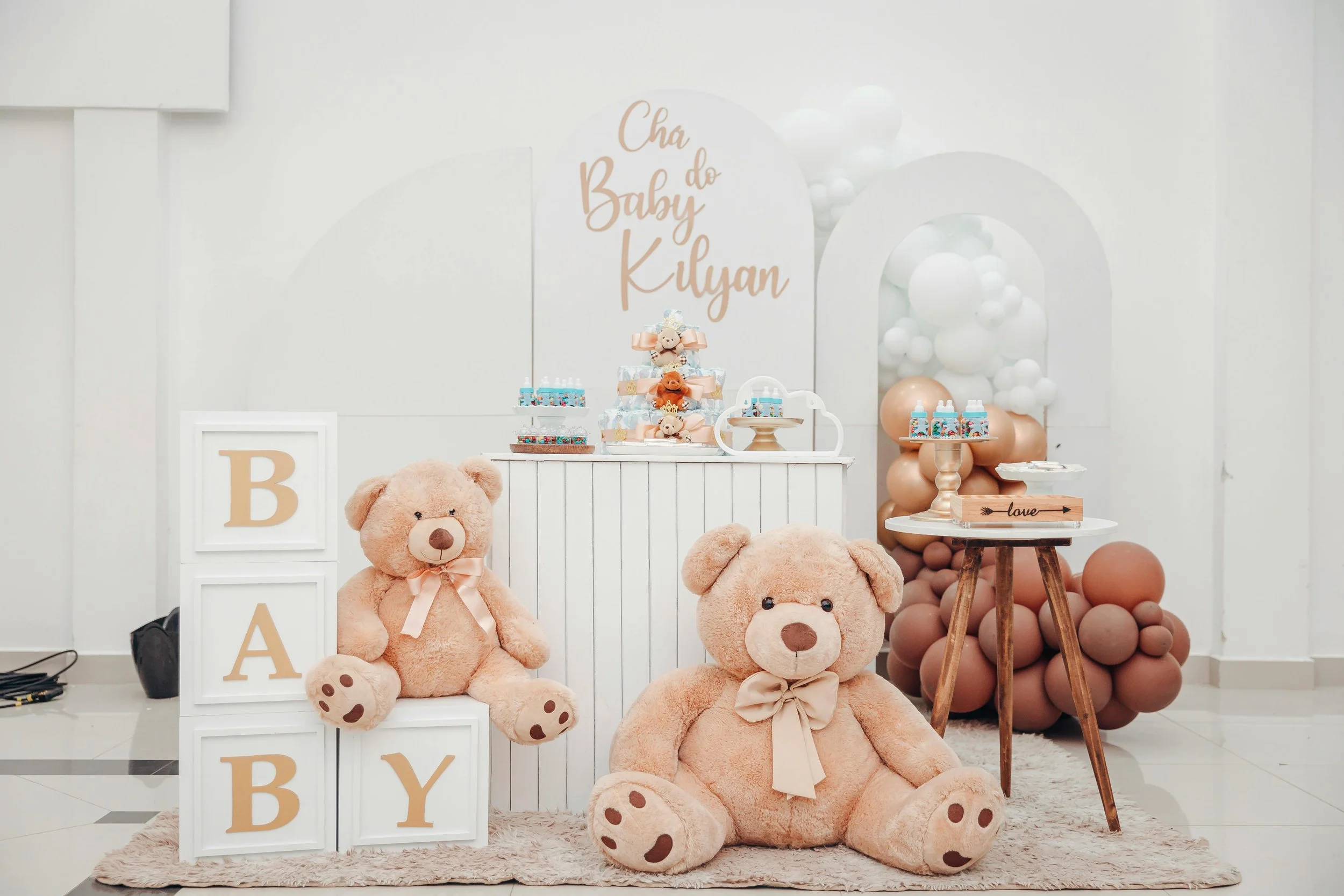 Decorations for a baby shower including plush teddy bears, balloon arrangements, wooden blocks with letters, and a dessert table with themed treats featuring bears.