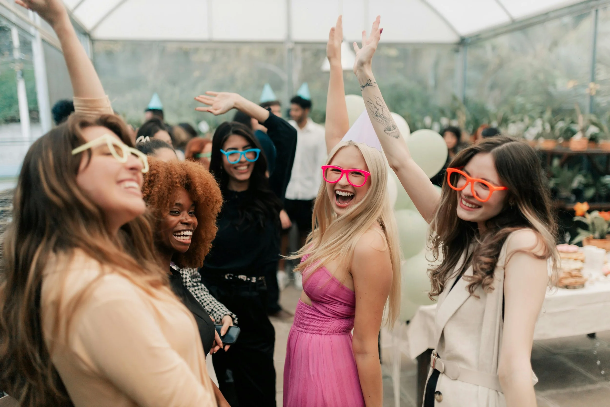 Group of diverse women celebrating at a party in a greenhouse with balloons and festive attire, wearing colorful glasses and smiling.