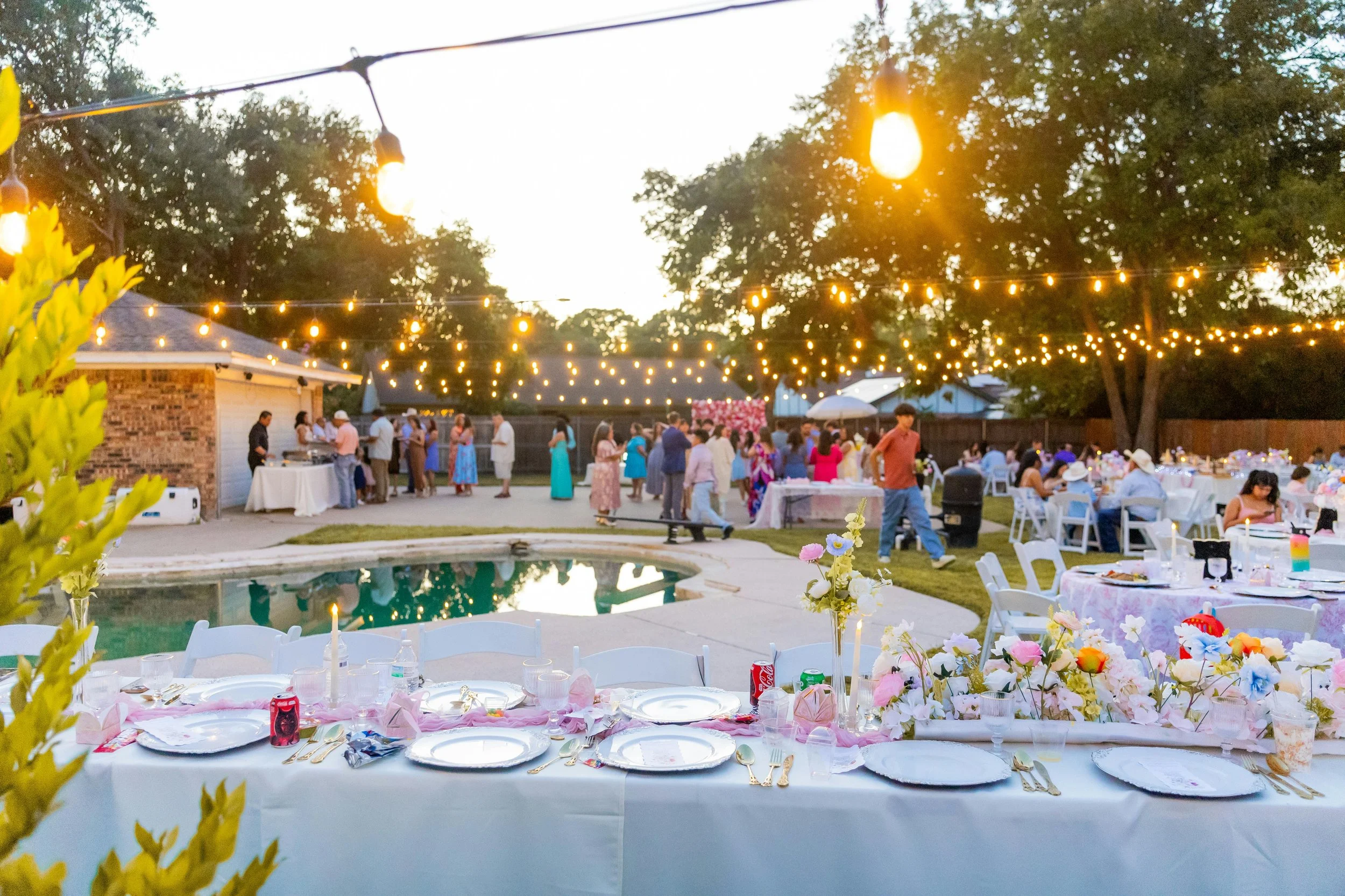 An outdoor party or celebration setup with tables, chairs, and string lights hanging above. People are gathered in the background, and there is a swimming pool in the foreground, surrounded by floral decorations and themed table settings.
