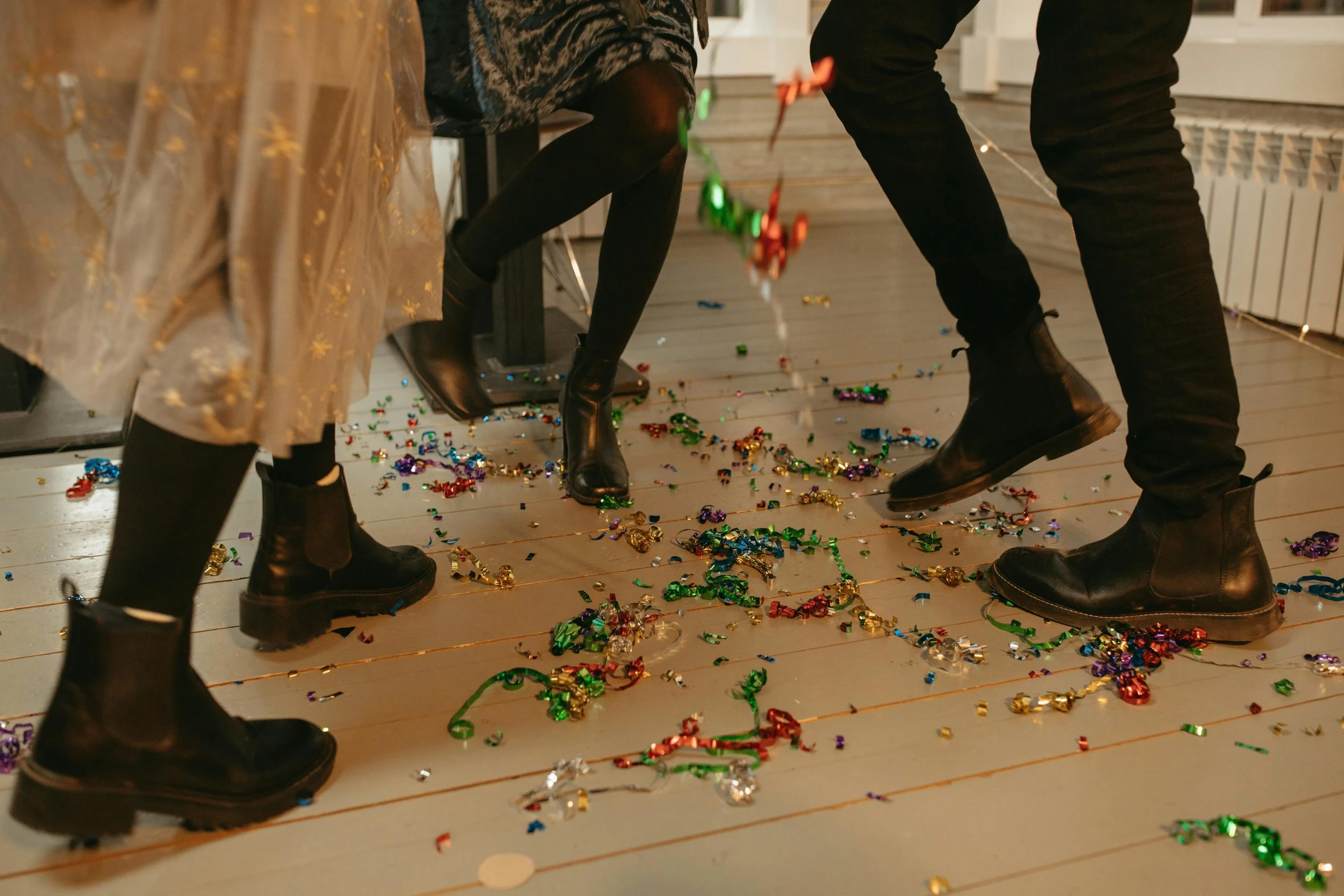 Close-up of people wearing black boots standing on a floor covered with colorful confetti, with some people reaching down, possibly at a celebration or party.