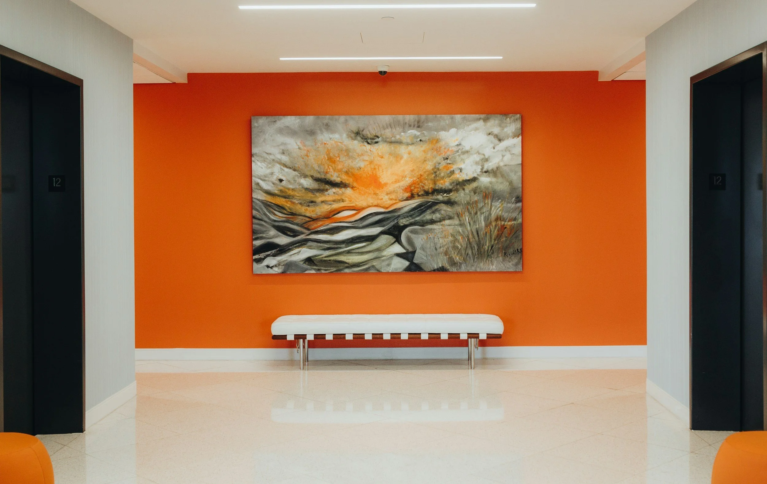 A modern hallway with an orange accent wall featuring an abstract painting, white bench, and two elevator doors.