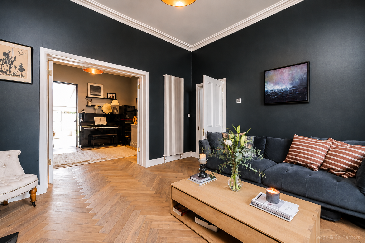Living room with a dark gray wall, a black sofa with striped pillows, a wooden coffee table with candles and a plant, and a framed painting hanging on the wall. An open door reveals a piano and a cozy room with decor and natural light.