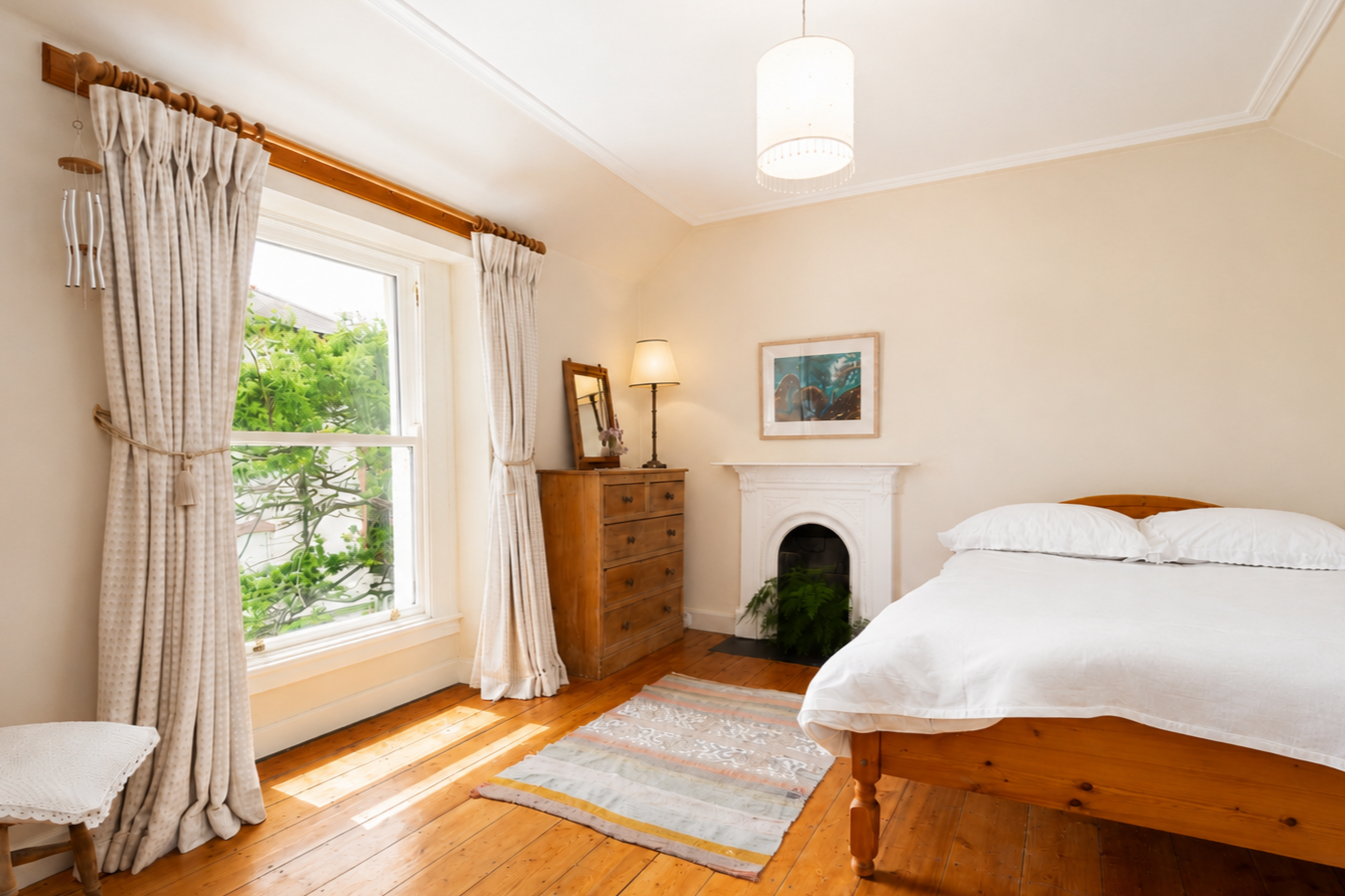 A cozy bedroom with a wooden bed, white bedding, wooden flooring, a window with beige curtains, a small rug, a wooden dresser with a mirror and lamp, a framed picture on the wall, a white fireplace with green plants inside, and a ceiling light fixture.