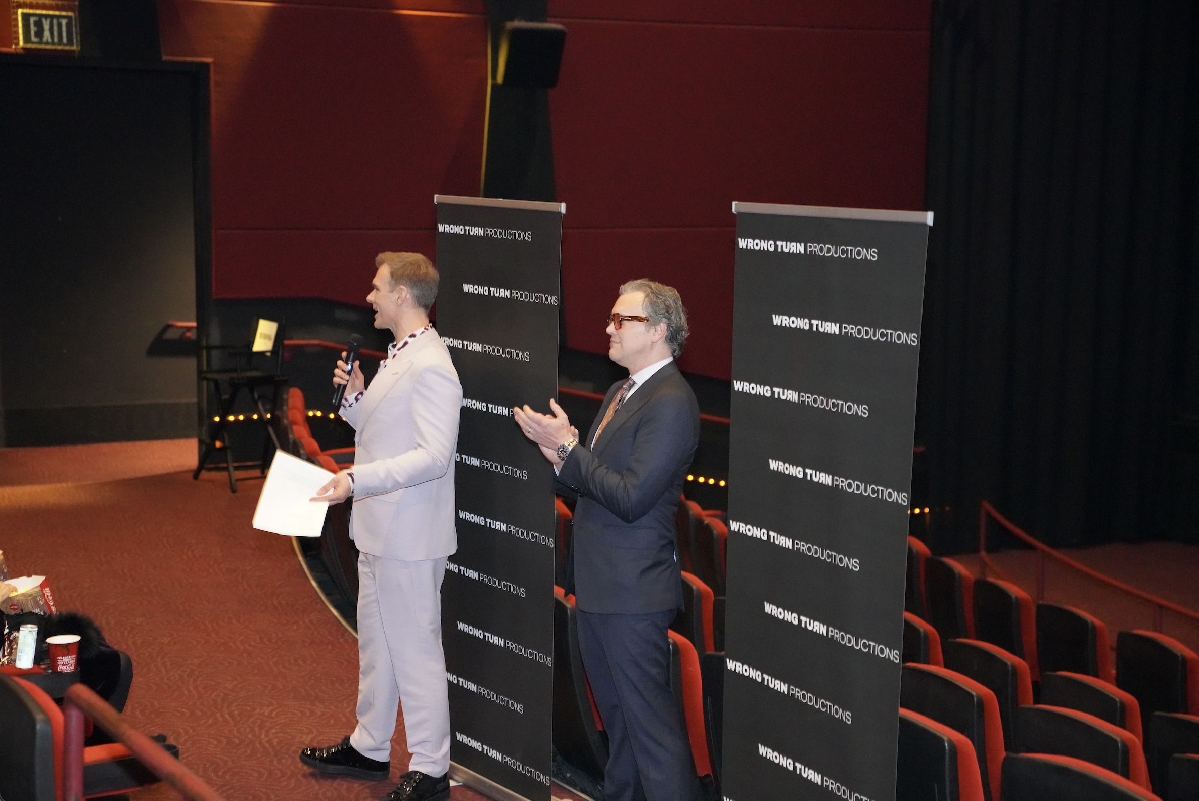 Two men are standing in front of black banners that say 'WRONG TURN PRODUCTIONS'. The man on the left is wearing a cream-colored suit with a patterned shirt and holding a microphone. The man on the right is wearing a dark suit with glasses. They are 