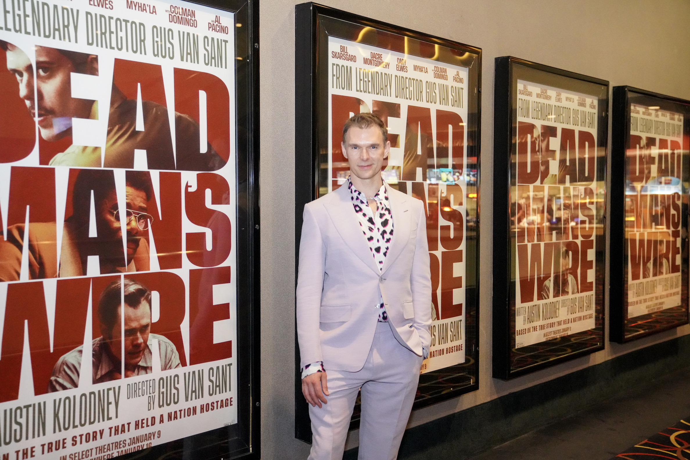 A young man with short light brown hair in a light-colored suit, standing in front of large movie posters for 'Fear'. The posters feature images of various characters and text about the movie.