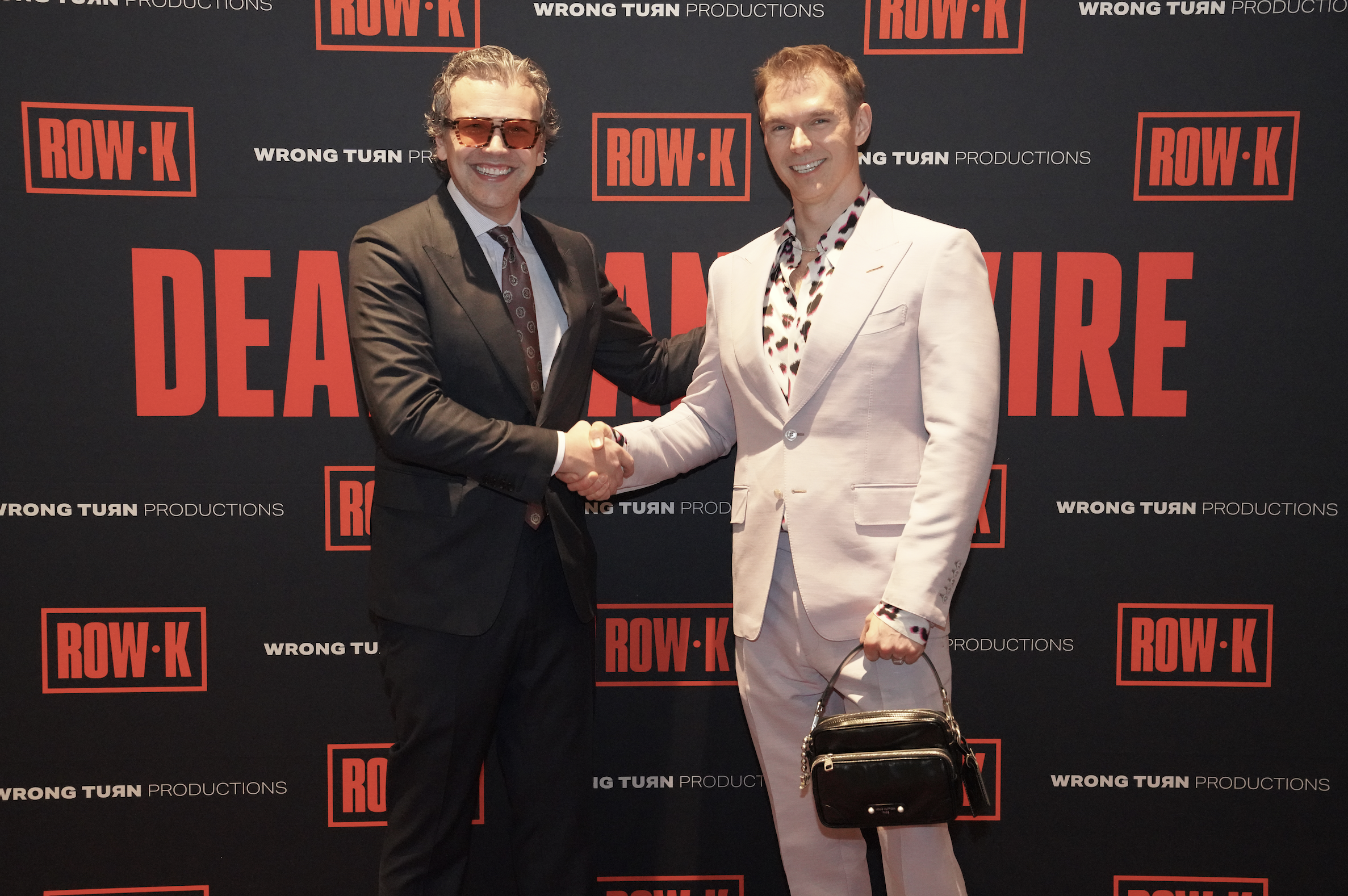 Two men shake hands and smile at camera in front of a black backdrop with red and white text and logos. One man wears a dark suit and sunglasses, the other a white suit with a patterned shirt.