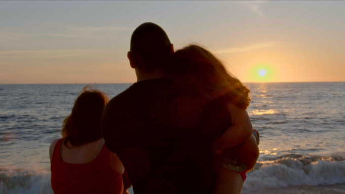 Three people, two adults and a child, watching sunset on the beach, standing close to the ocean.