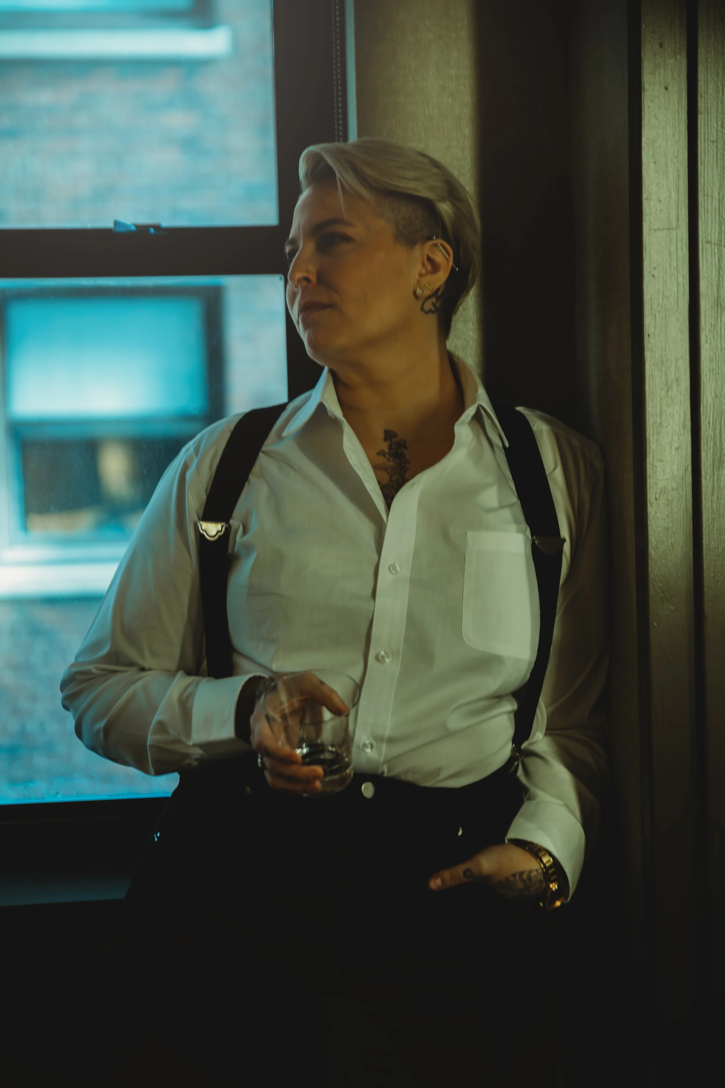 A person with short, styled blonde hair, wearing a white button-down shirt with a pocket, black suspenders, and multiple earrings, stands by a window holding a glass of drink. They have tattoos visible on their neck and hand, and are looking to the side with a serious expression.
