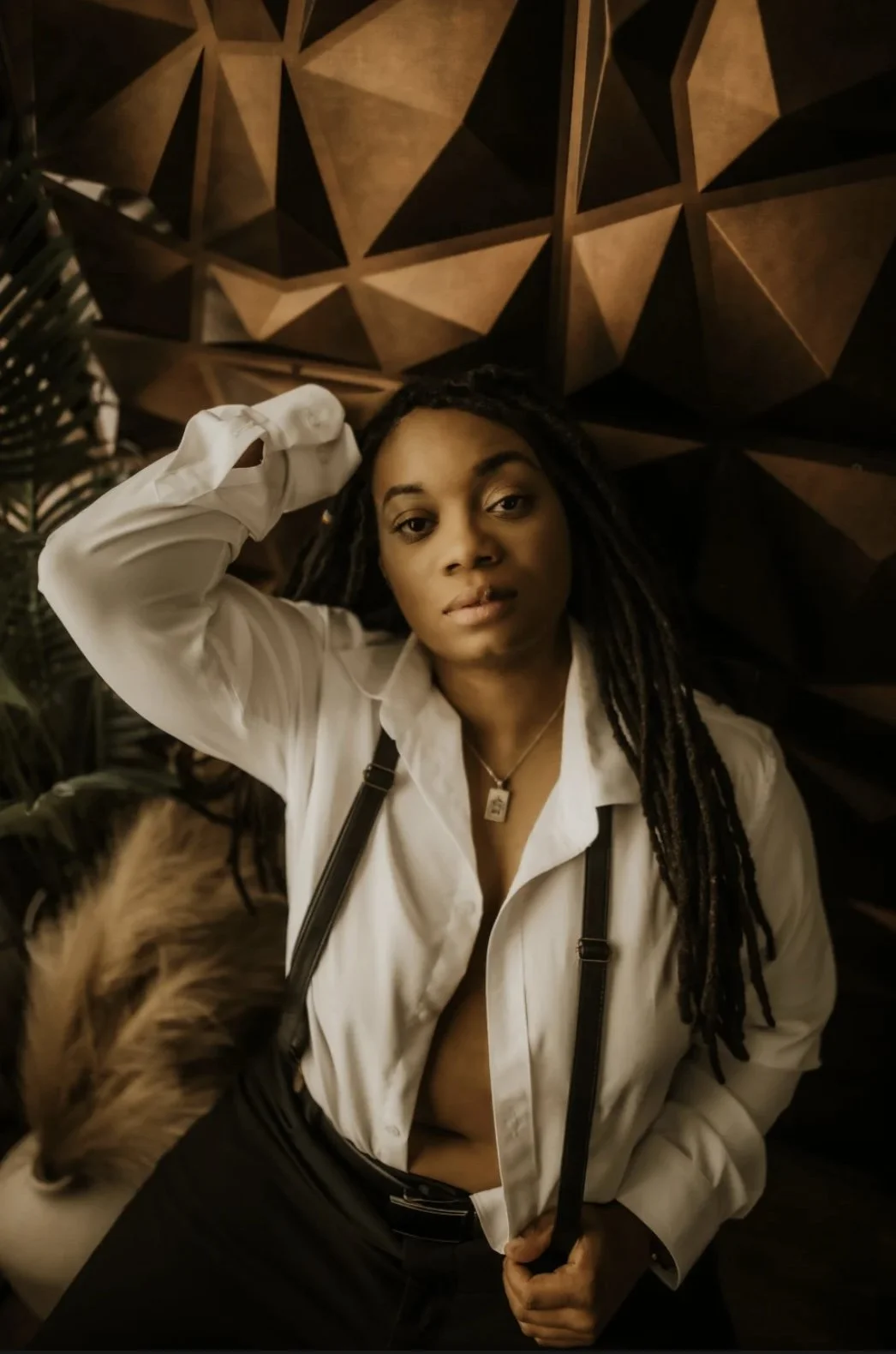 A woman with long braided hair, wearing a white shirt and black pants, is sitting against a geometric wooden wall. She has a necklace and is holding what appears to be a strap or belt. A dog is partially visible at the lower left corner of the image.