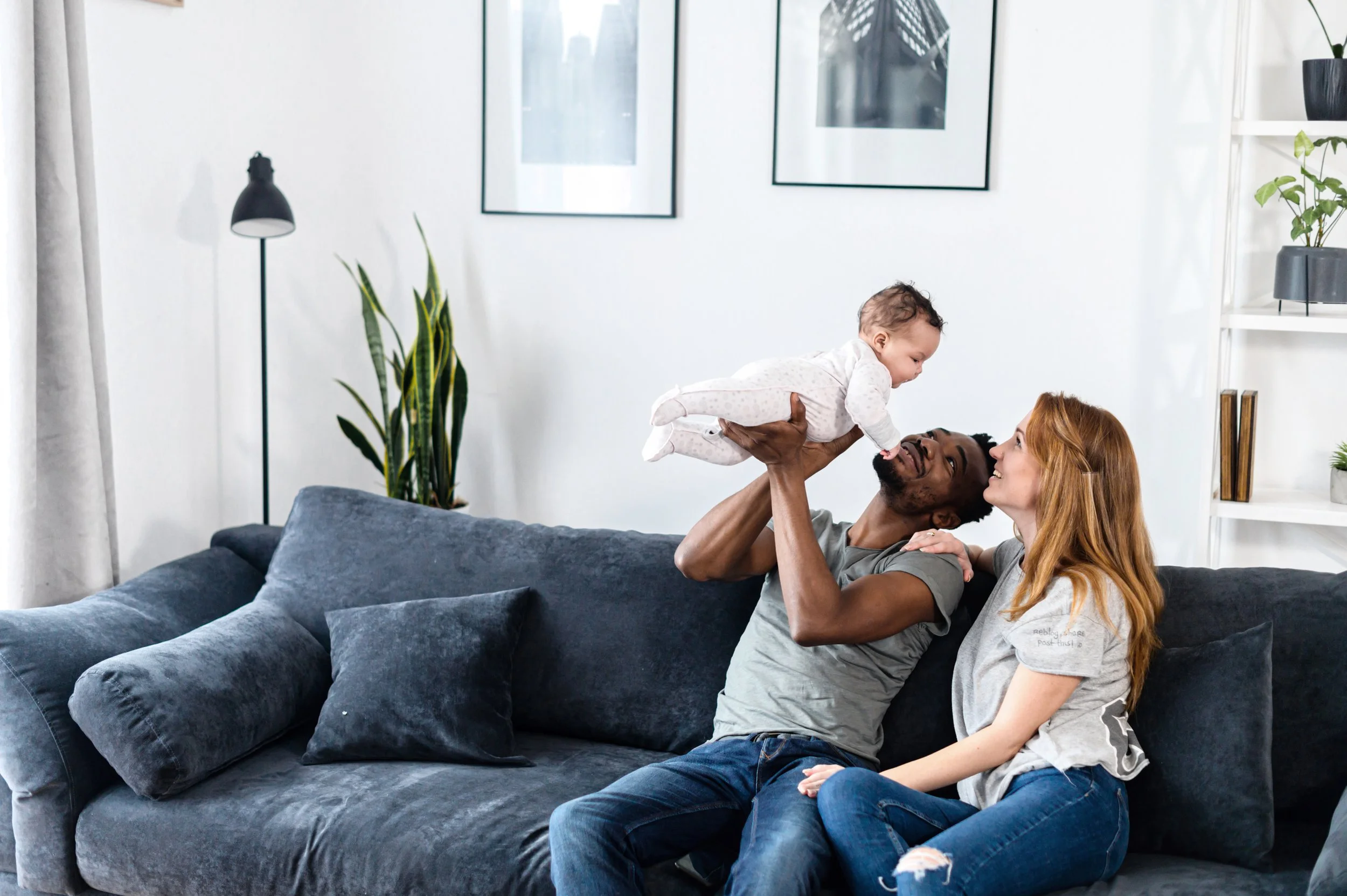 A family of three, a man, a woman, and a baby, enjoying playtime together in a living room. The man lifts the baby into the air while lying on a dark gray couch, and the woman sits nearby smiling.