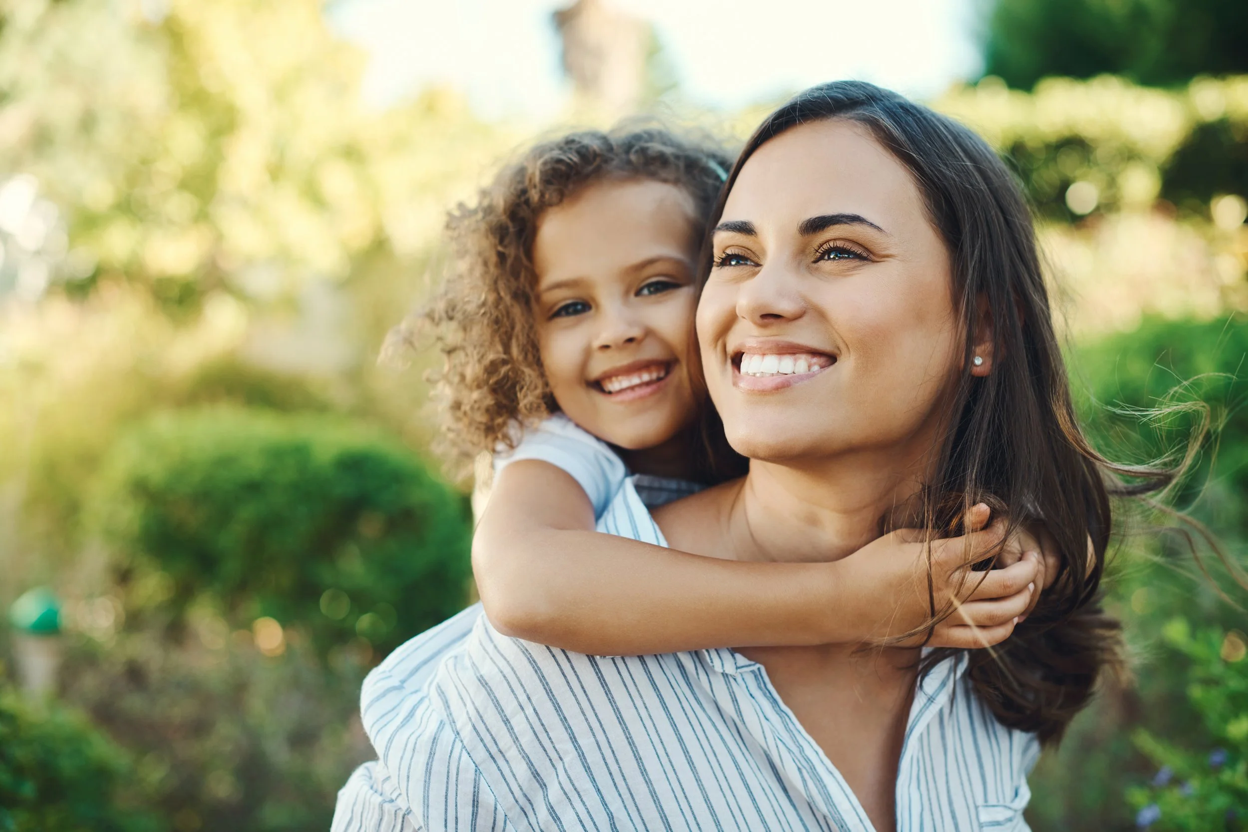 A woman with long dark hair and a young girl with curly hair smiling outdoors in a park, with sunlight filtering through trees in the background.