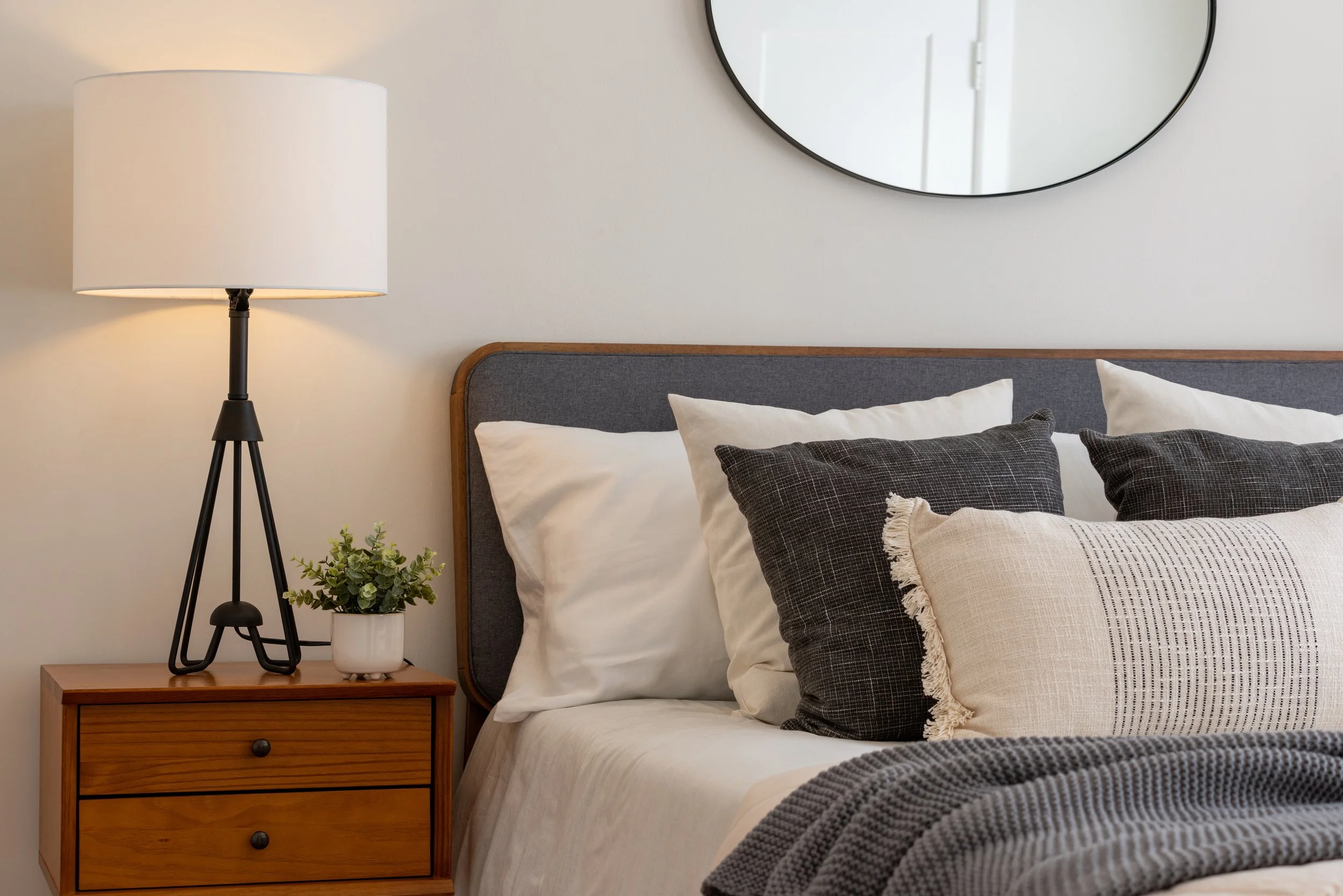 A cozy bedroom with a neatly made bed, decorative pillows, a wooden bedside table with a plant, and a tall lamp beside the bed, with a round mirror on the wall.