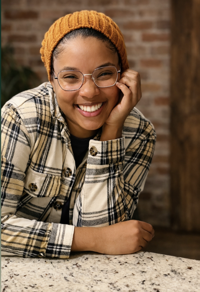 A smiling young woman wearing glasses, an orange knit beanie, and a plaid jacket sitting at a speckled countertop, with a brick wall in the background.