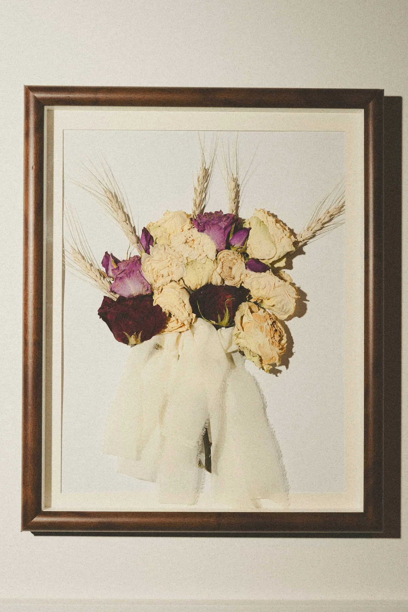 Framed art of dried flower bouquet, including roses and wheat, hanging on a beige wall.