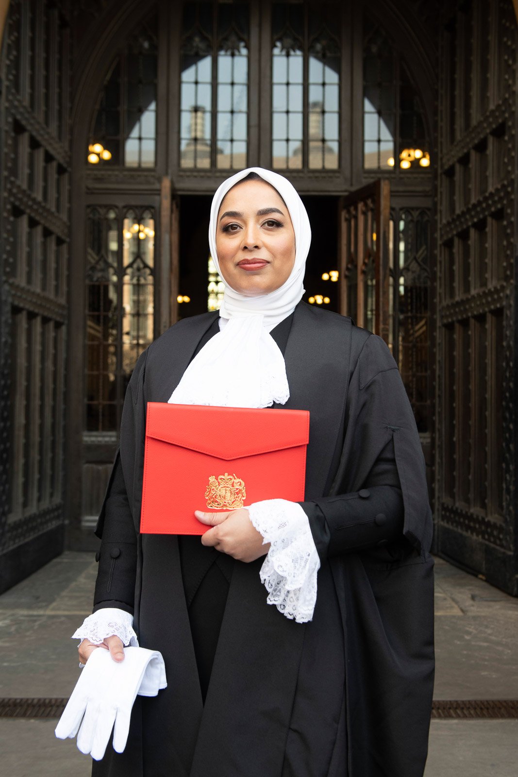 A woman in a kings counsel robe and white hijab holding a letter patent from Her Majesty Queen Elizabeth II and white gloves in front of a historic building.
