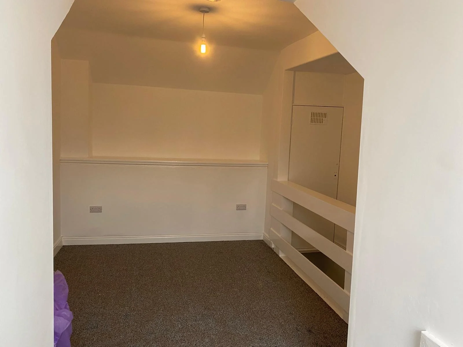 Empty small room with sloped ceiling, beige walls, brown carpet, a single hanging light bulb, and white built-in shelving or cabinetry.
