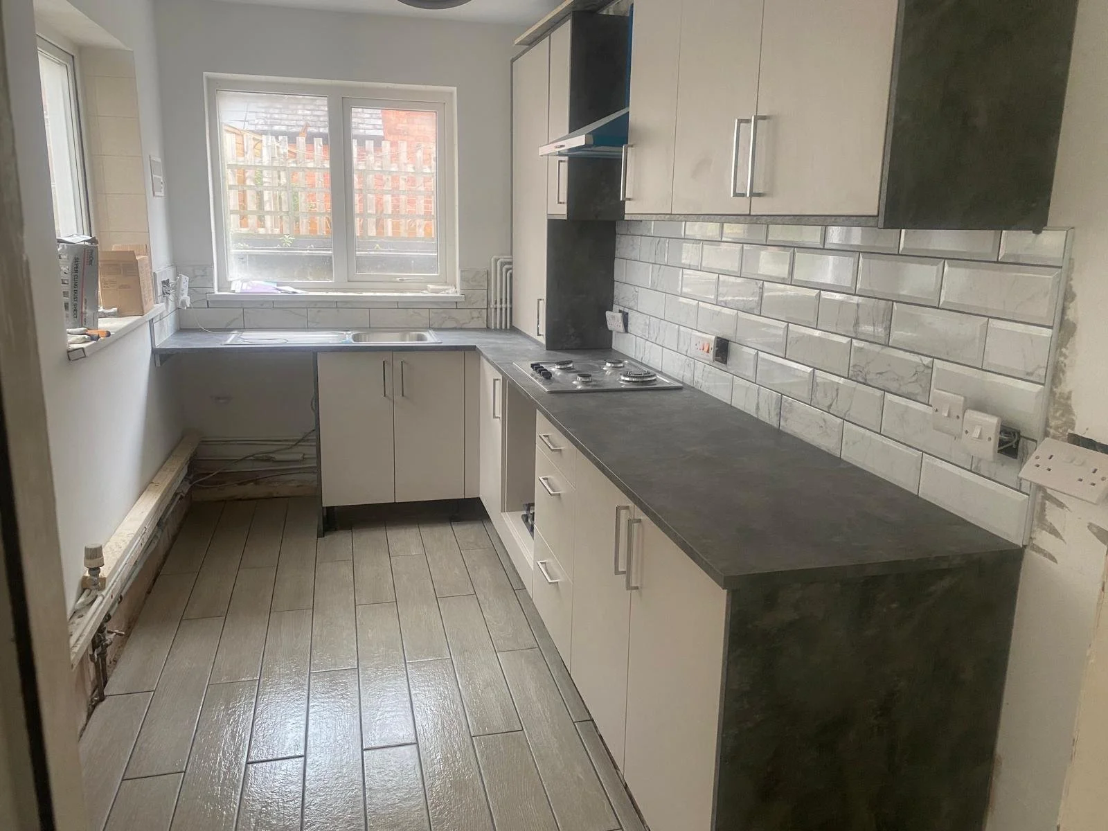 Unfinished kitchen with white cabinets, grey countertops, a white tiled backsplash, and a window above the sink, with some construction materials and wires visible.