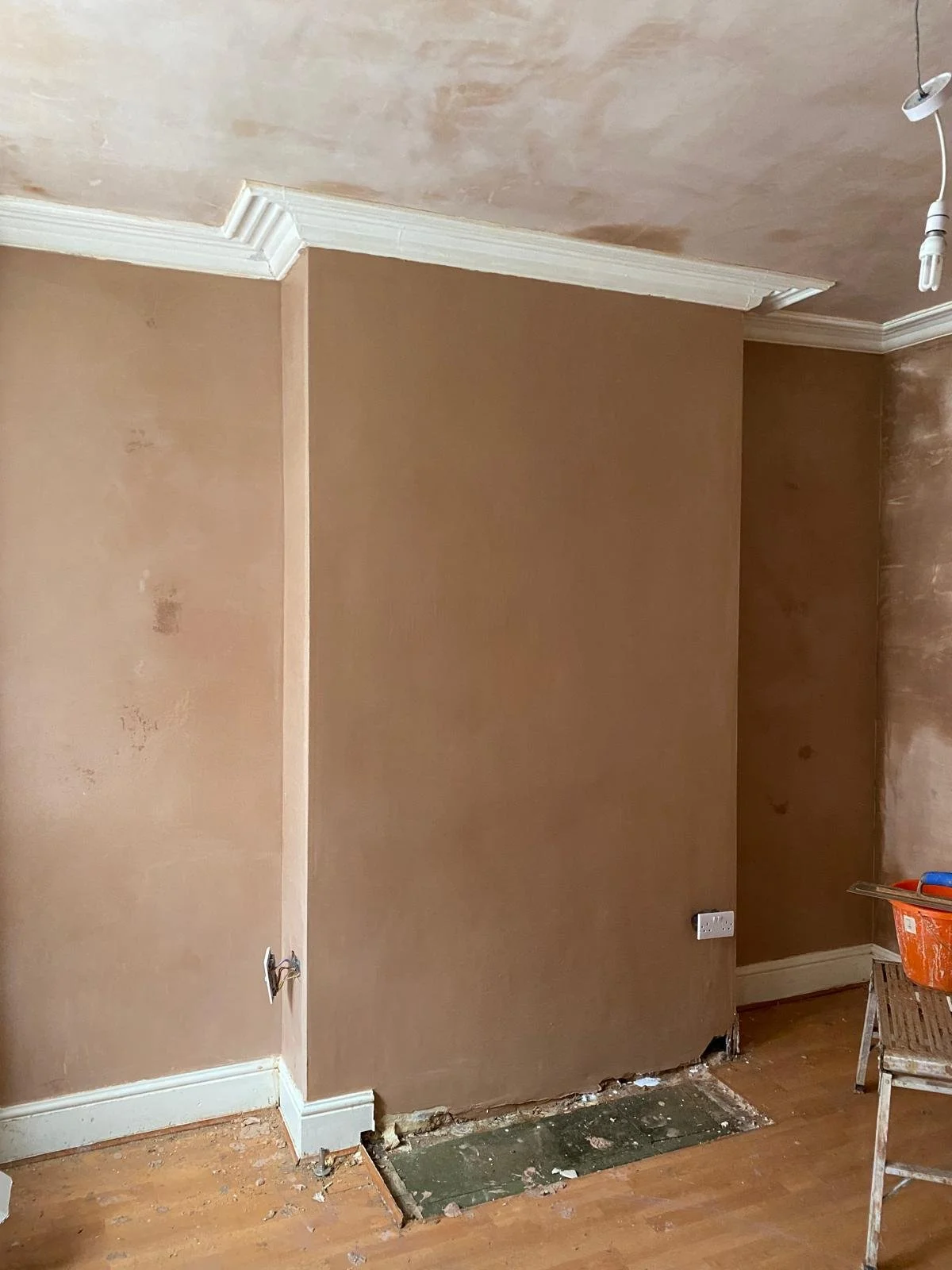 A room undergoing renovation with beige painted walls, crown molding, exposed electrical outlets, and an open space in the floor likely for plumbing or electrical work, with a step ladder and tools nearby.