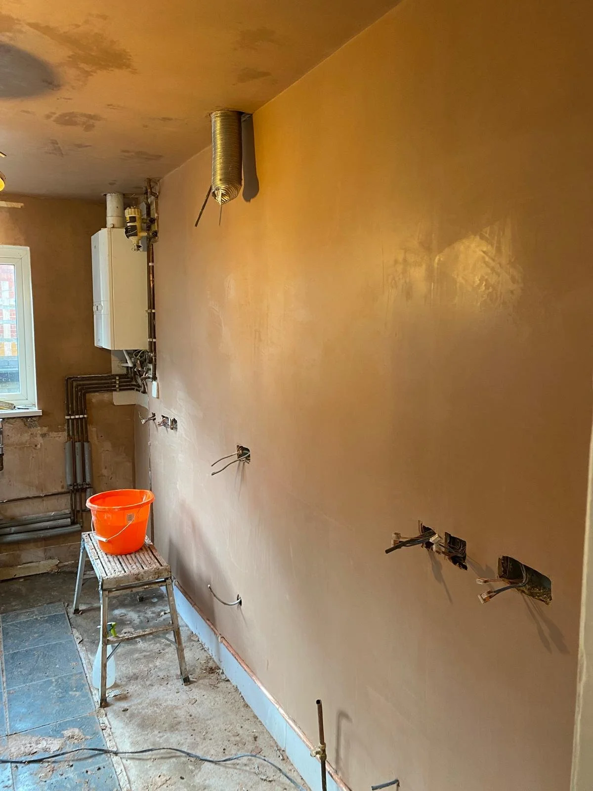 Interior of a room under construction or renovation, with a partially finished wall, exposed pipes, and electrical wiring, a small step ladder with an orange bucket on top, and a window in the background.