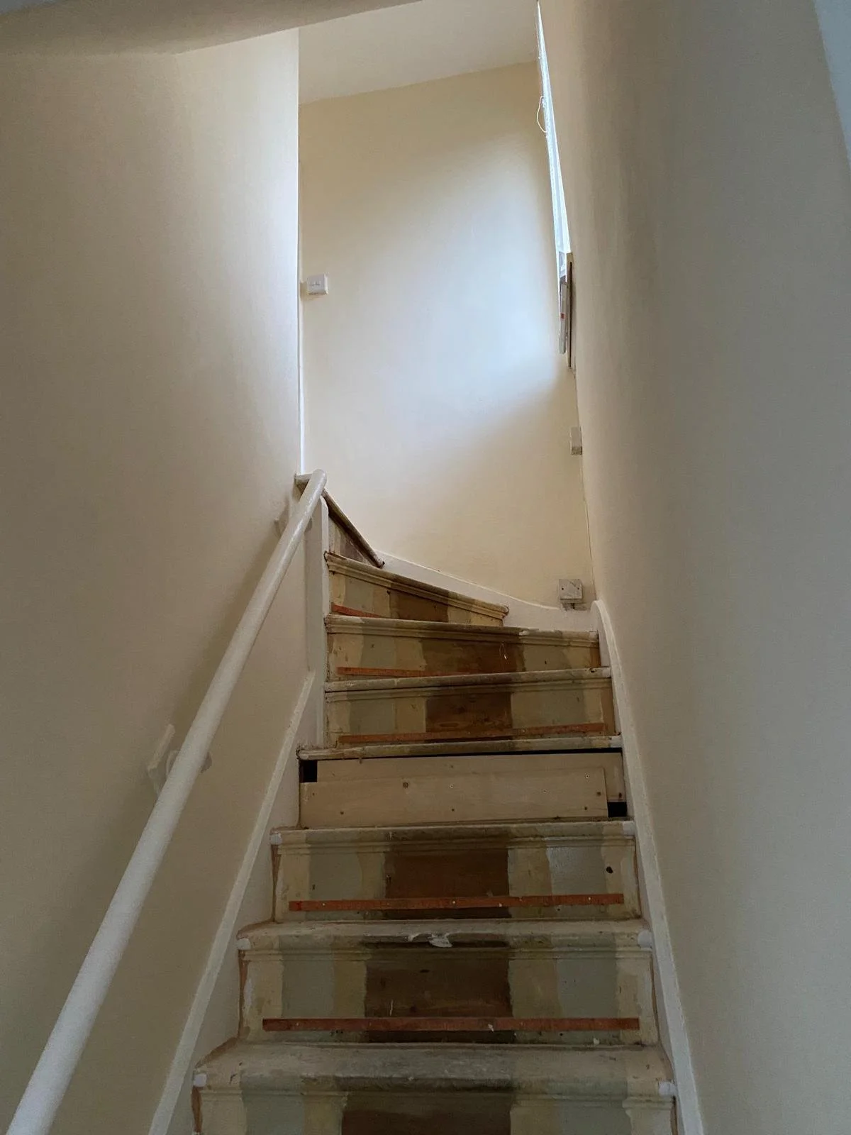 Interior view of a staircase under renovation, showing unfinished wooden steps and a white handrail on the left side.