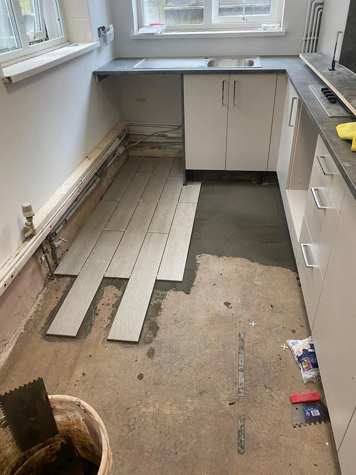 Kitchen under renovation with partially installed light-colored floor tiles, a black underlayment layer, white cabinets, a sink, window, and construction tools and materials scattered around.