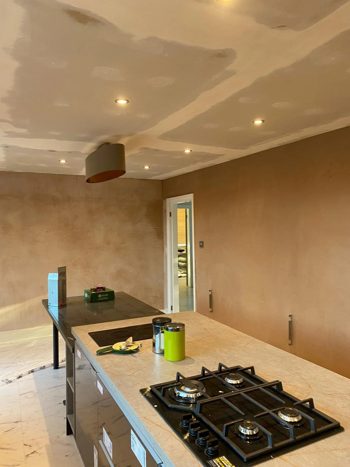 A kitchen under renovation with a new ceiling with recessed lights and a ceiling fixture, an island with a cooktop, and wall textures in progress.